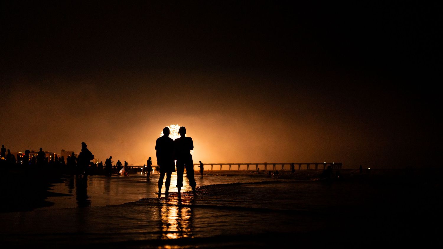 Two silhouetted figures stand on a beach at night, illuminated by a warm golden glow on the horizon near a pier.