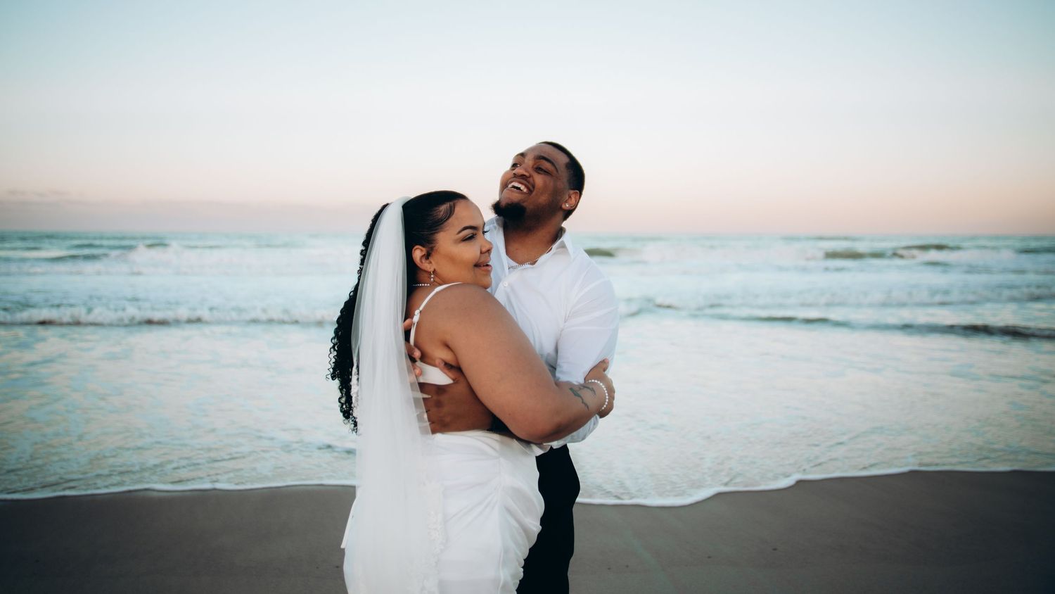 Newlywed couple embracing on a beach at sunset, bride in white dress and veil, groom in white shirt.