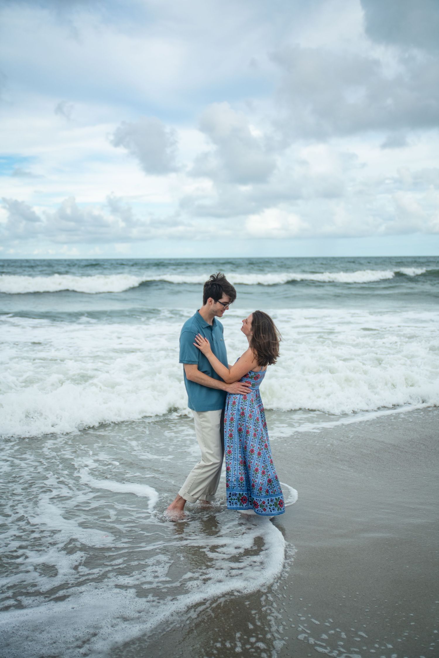 A couple embraces in the ocean waves as they crash around their feet on a cloudy day.