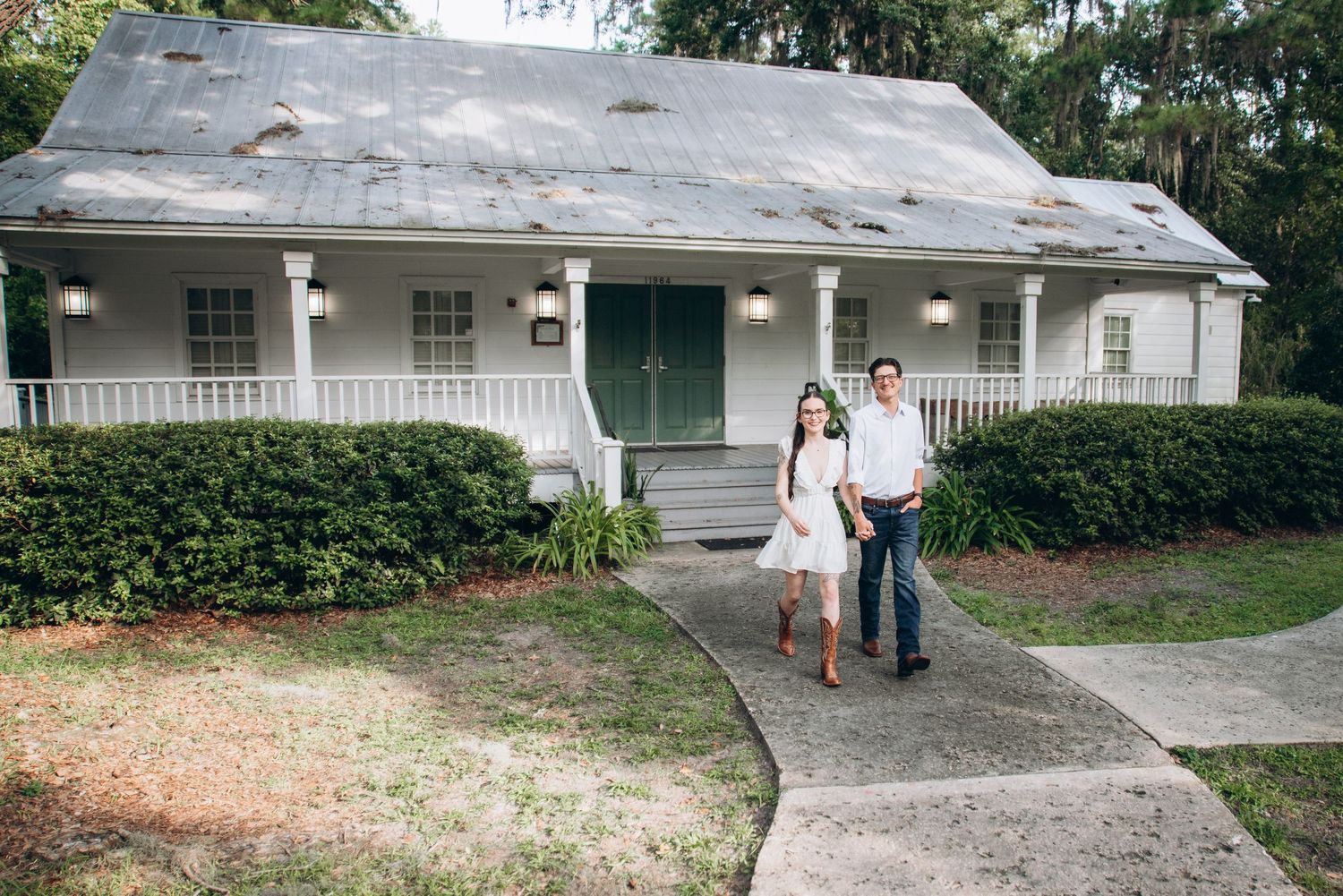 White farmhouse with wraparound porch and green shutters sits nestled among trees on a sunny day.