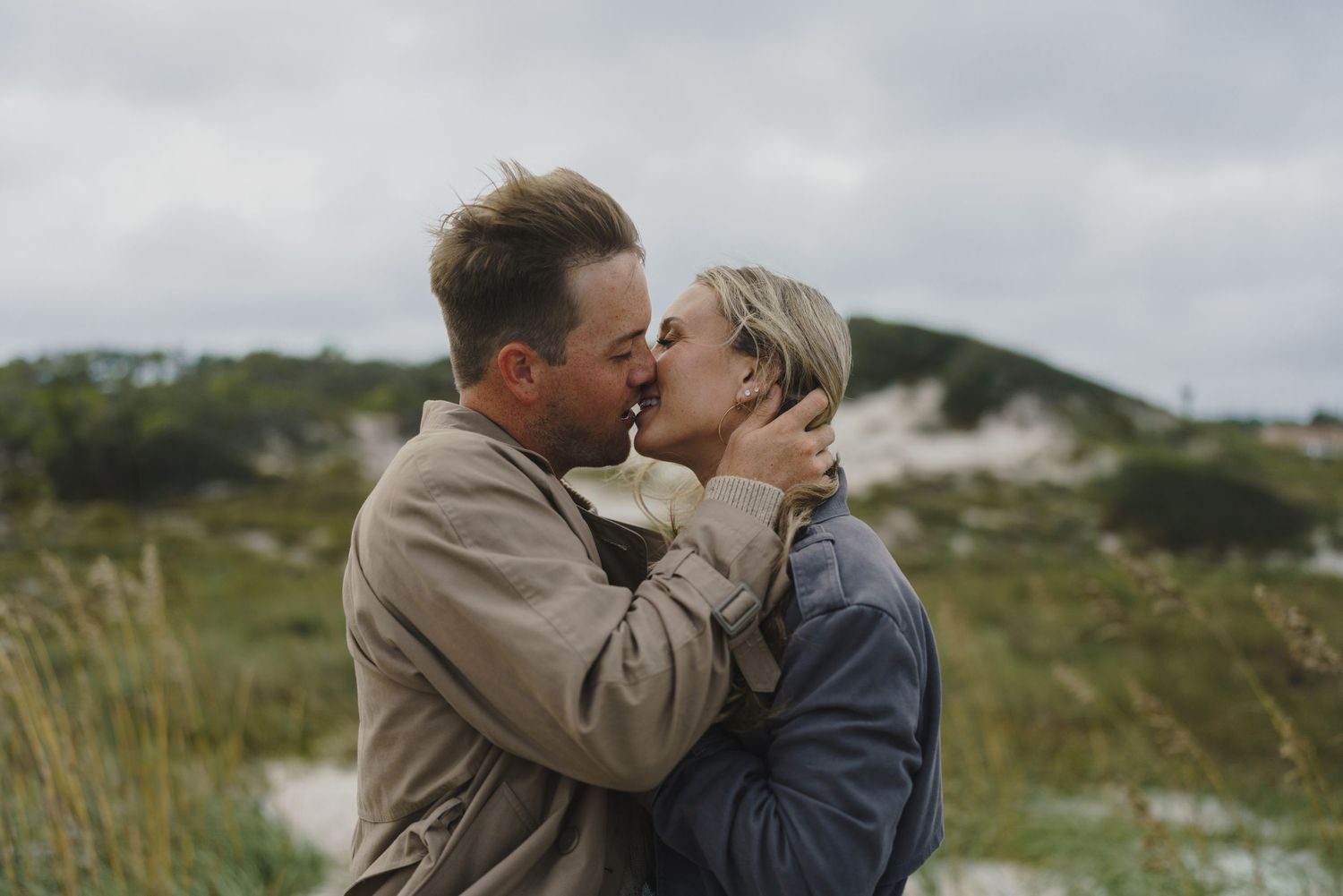 A romantic couple embracing in a scenic coastal landscape with grassy dunes.