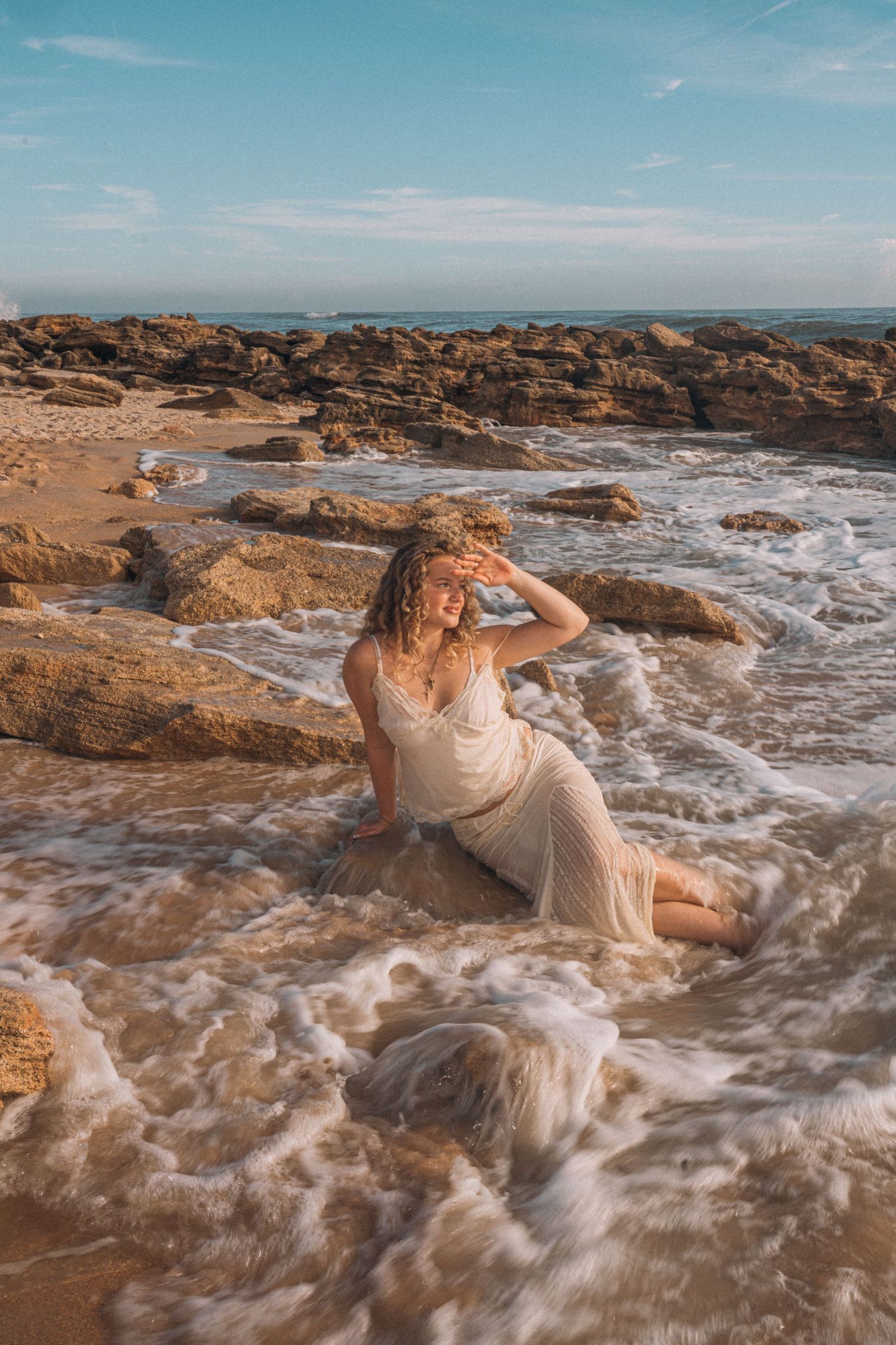 Woman in white dress sitting in ocean waves on rocky beach, smiling with hand raised, golden hour light.