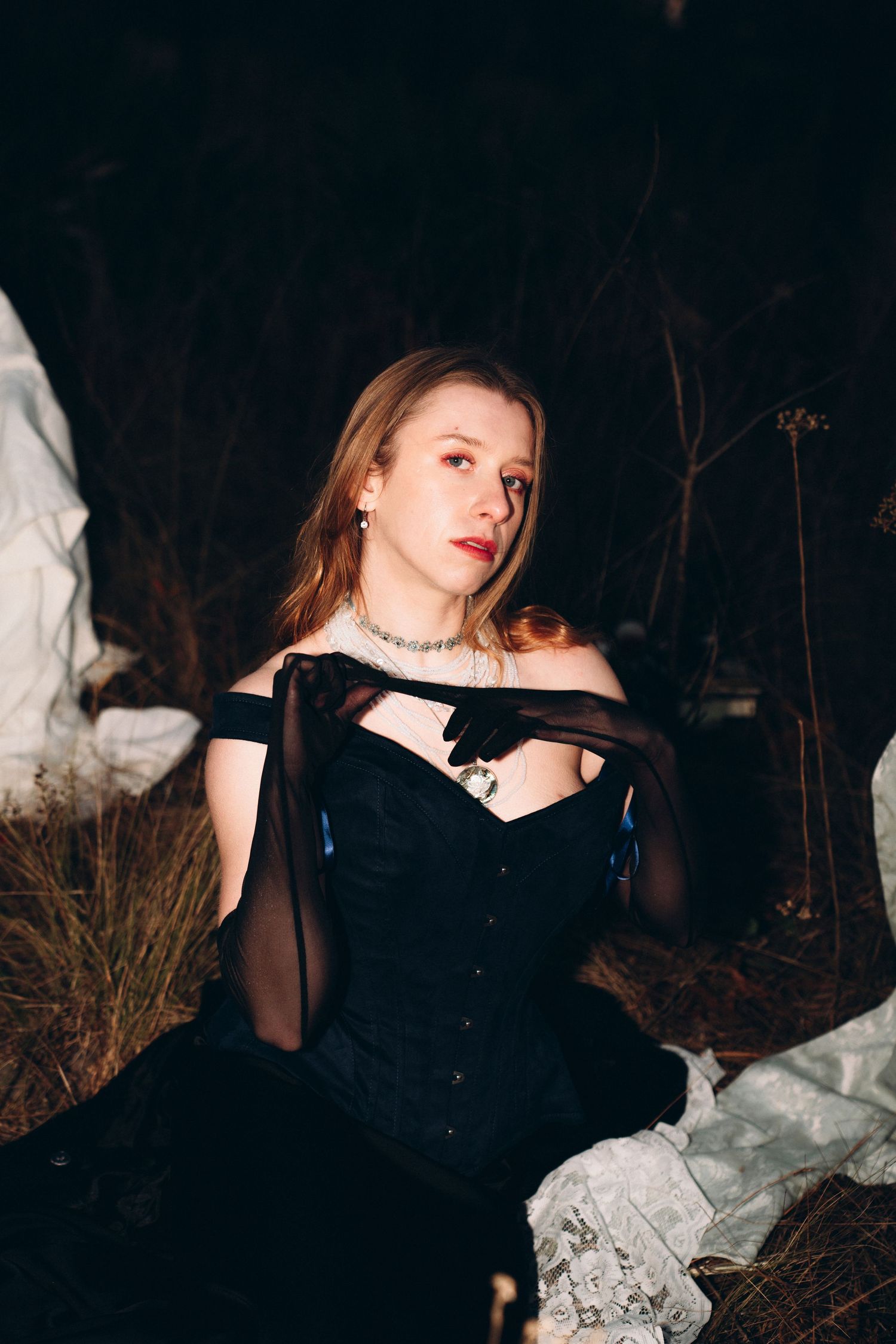 Young woman in black corset gown with sheer gloves and silver necklace sitting outdoors at night.
