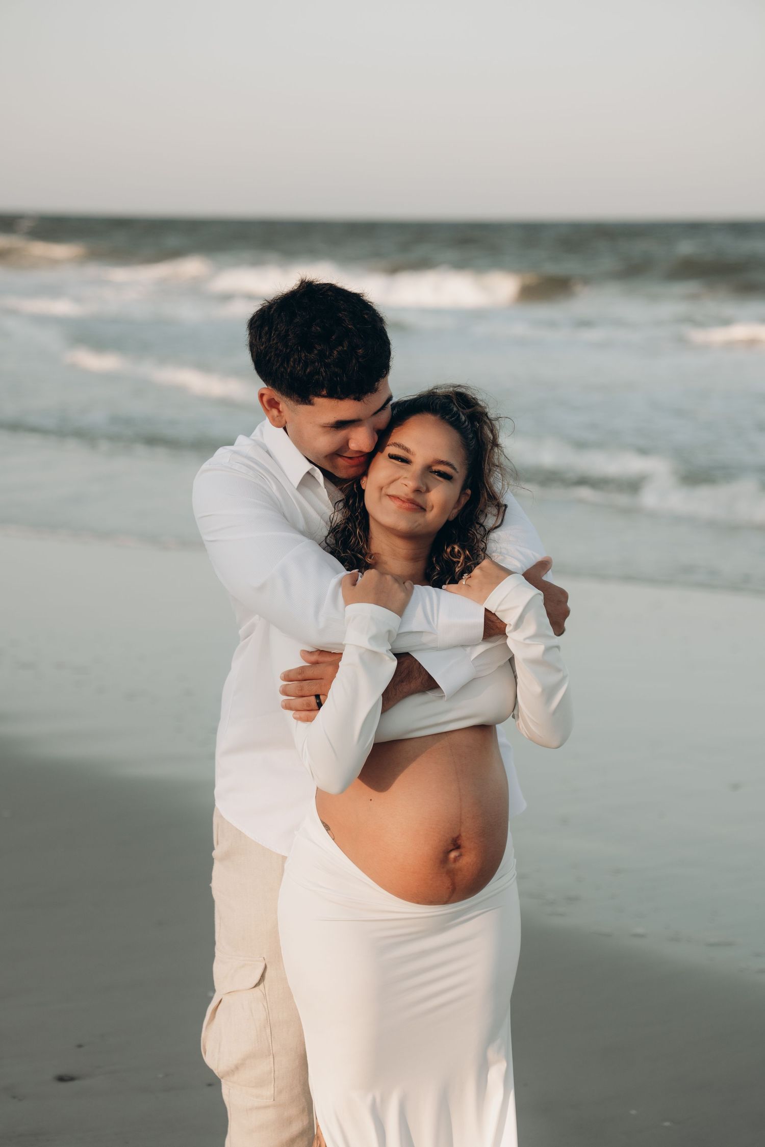 A romantic beach photoshoot with a couple in white outfits embracing by the ocean waves.