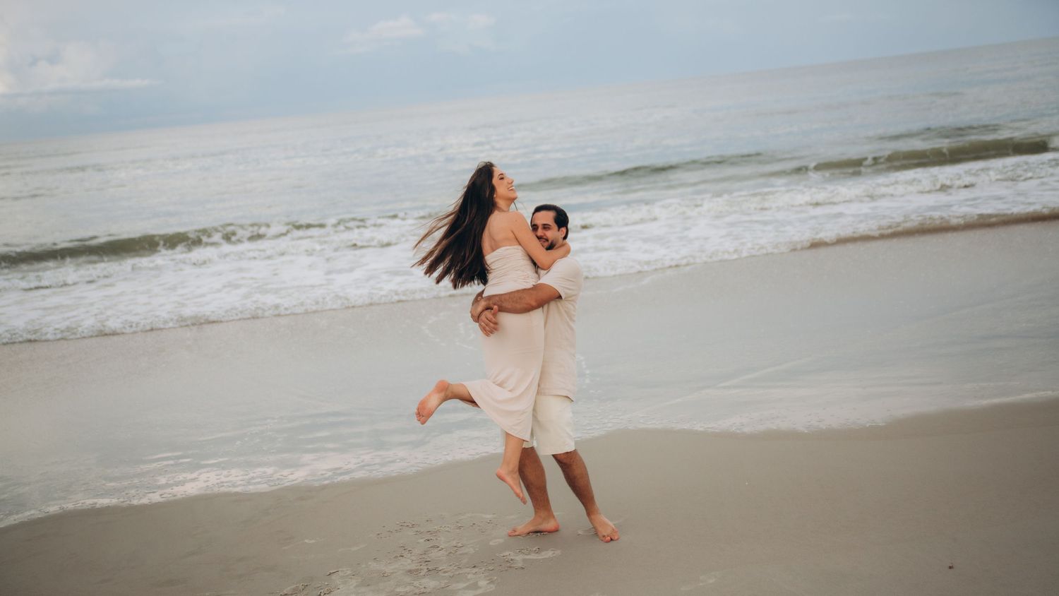 A couple in white outfits walking and playing together on a sandy beach at sunset.