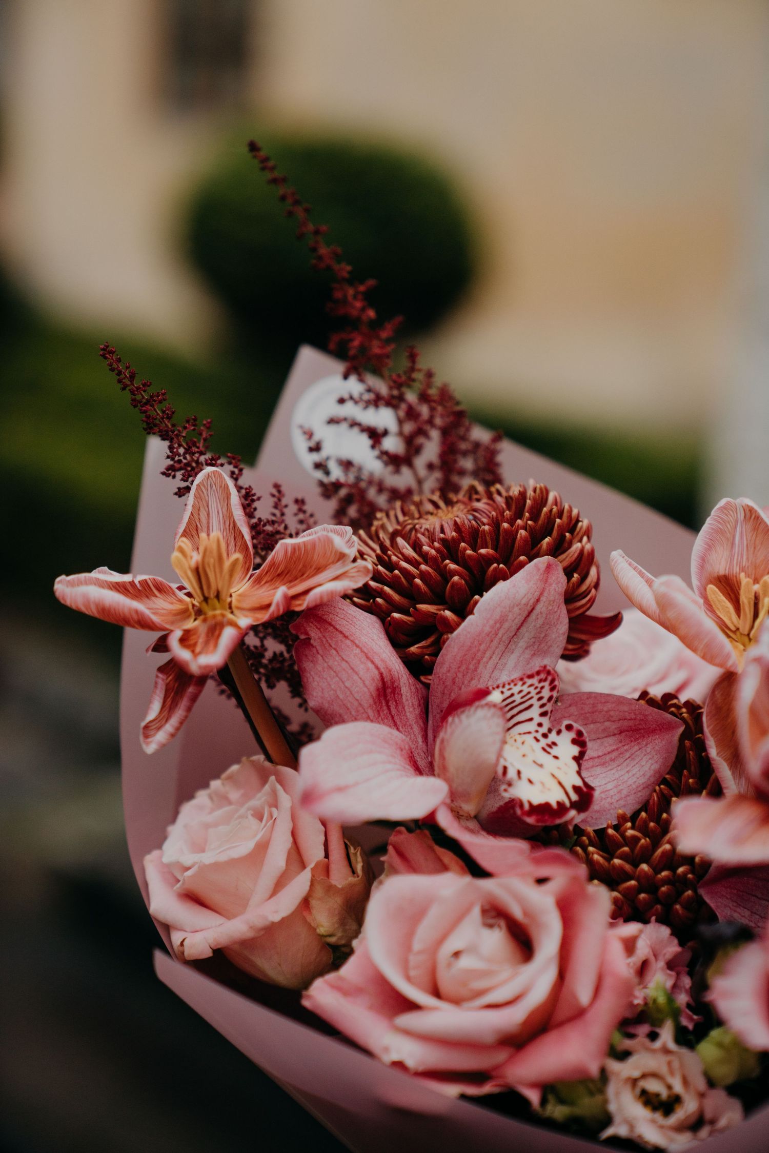 A lush pink bouquet featuring roses, orchids, and chrysanthemums wrapped in pink paper, set against a blurred garden background.