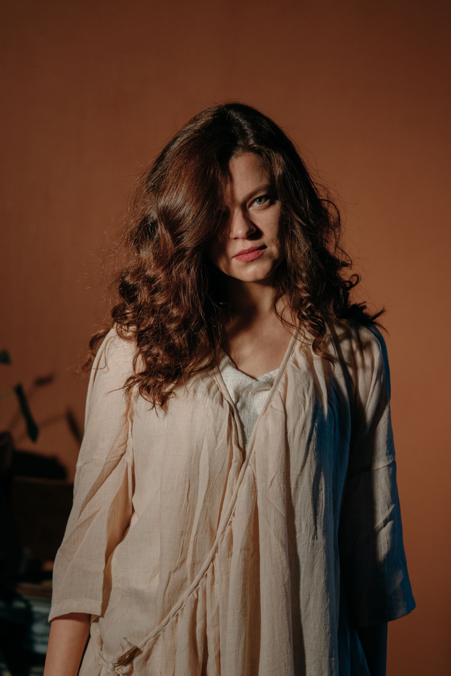 Young woman with curly brown hair wearing a cream linen dress, posing against a warm brown background.