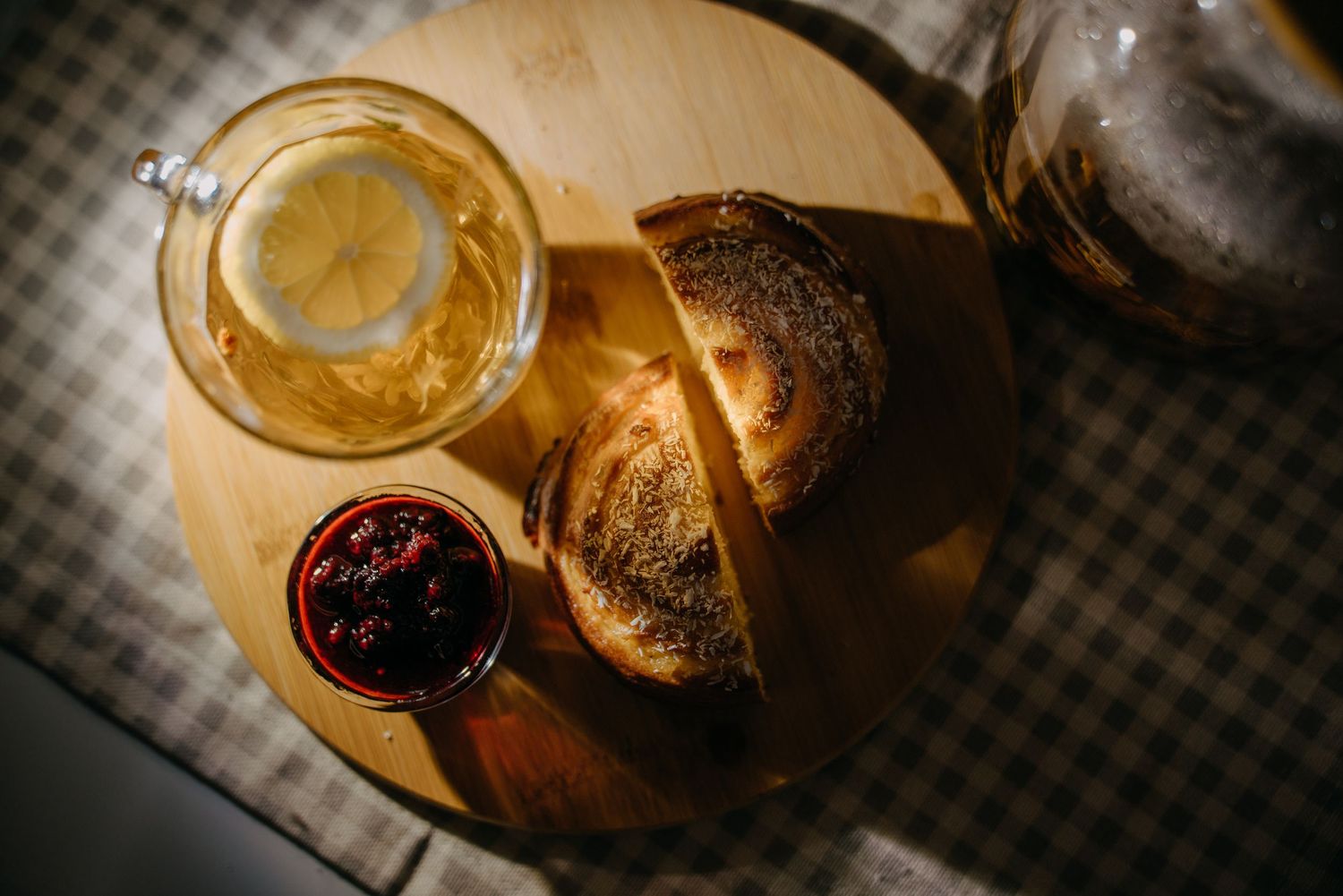 Overhead view of toasted bread, raspberry jam, and lemon tea on a wooden board with checkered cloth.