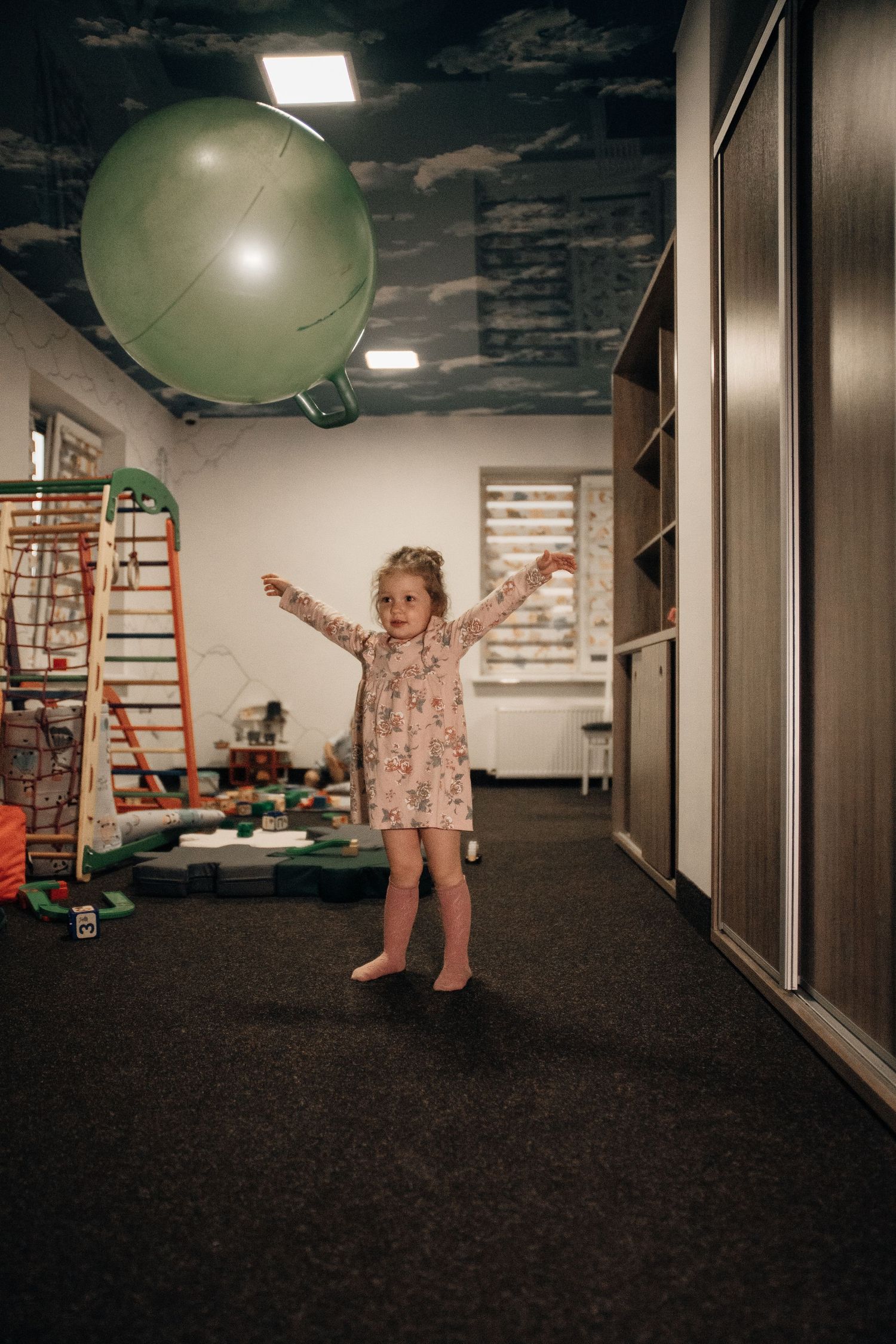 Young girl in floral dress playing with a green balloon in a decorated children's playroom with toys.