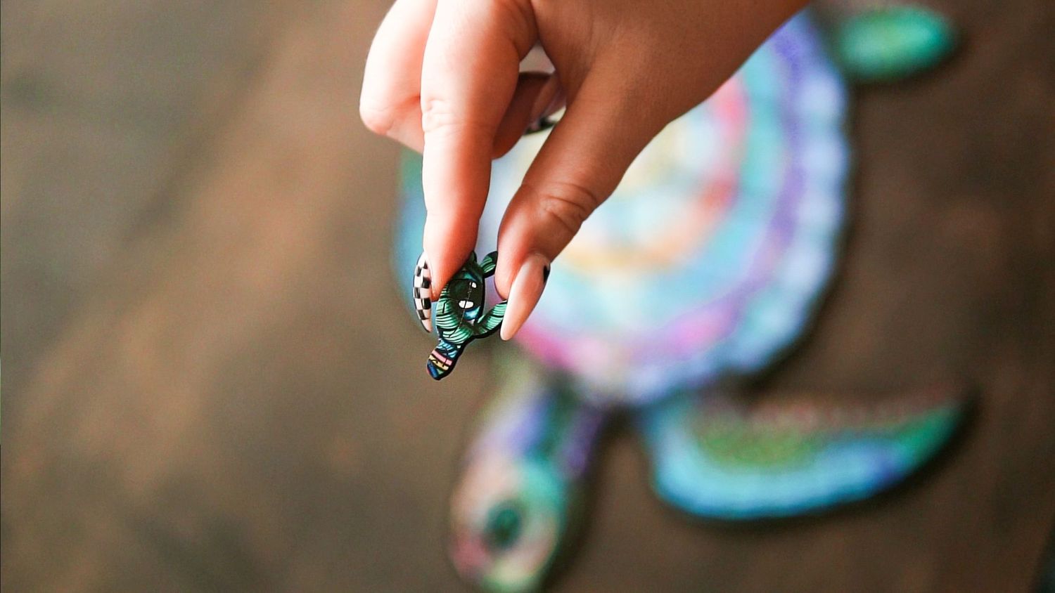 Hand holding a small decorative turtle figurine over a colorful glitter turtle art piece on a wooden surface.