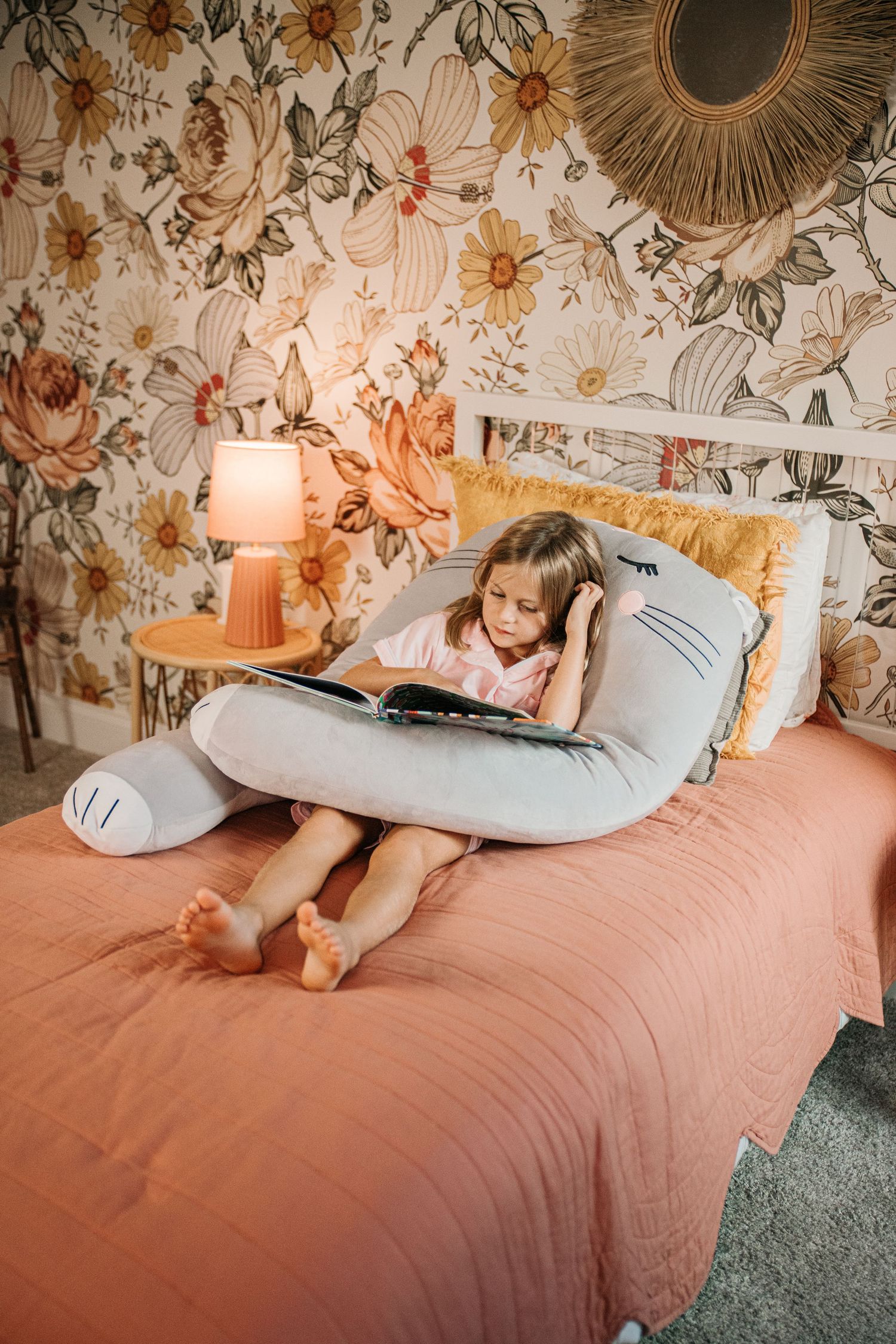 Young girl reading a book on a pink bed with floral wallpaper, cozy lamp, and a large shark pillow.