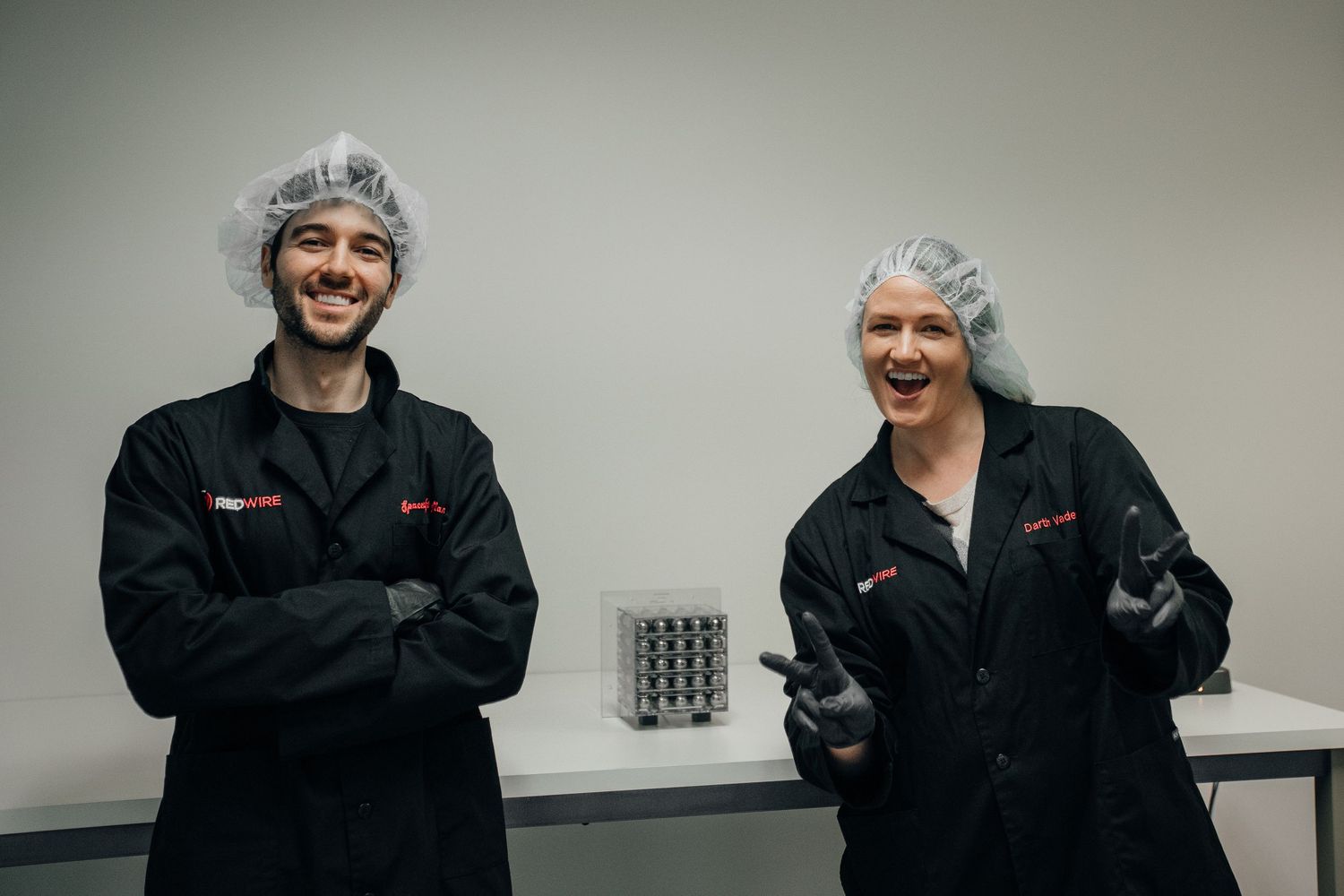 Two food industry workers in black uniforms and hair nets smiling in a clean production facility.