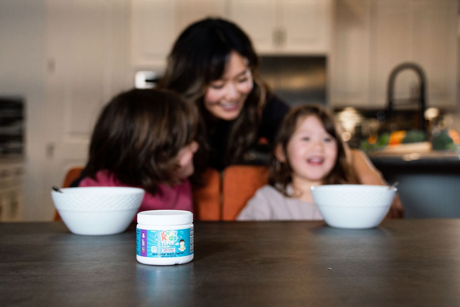 Mom laughing with two children over bowls, supplement jar in foreground.