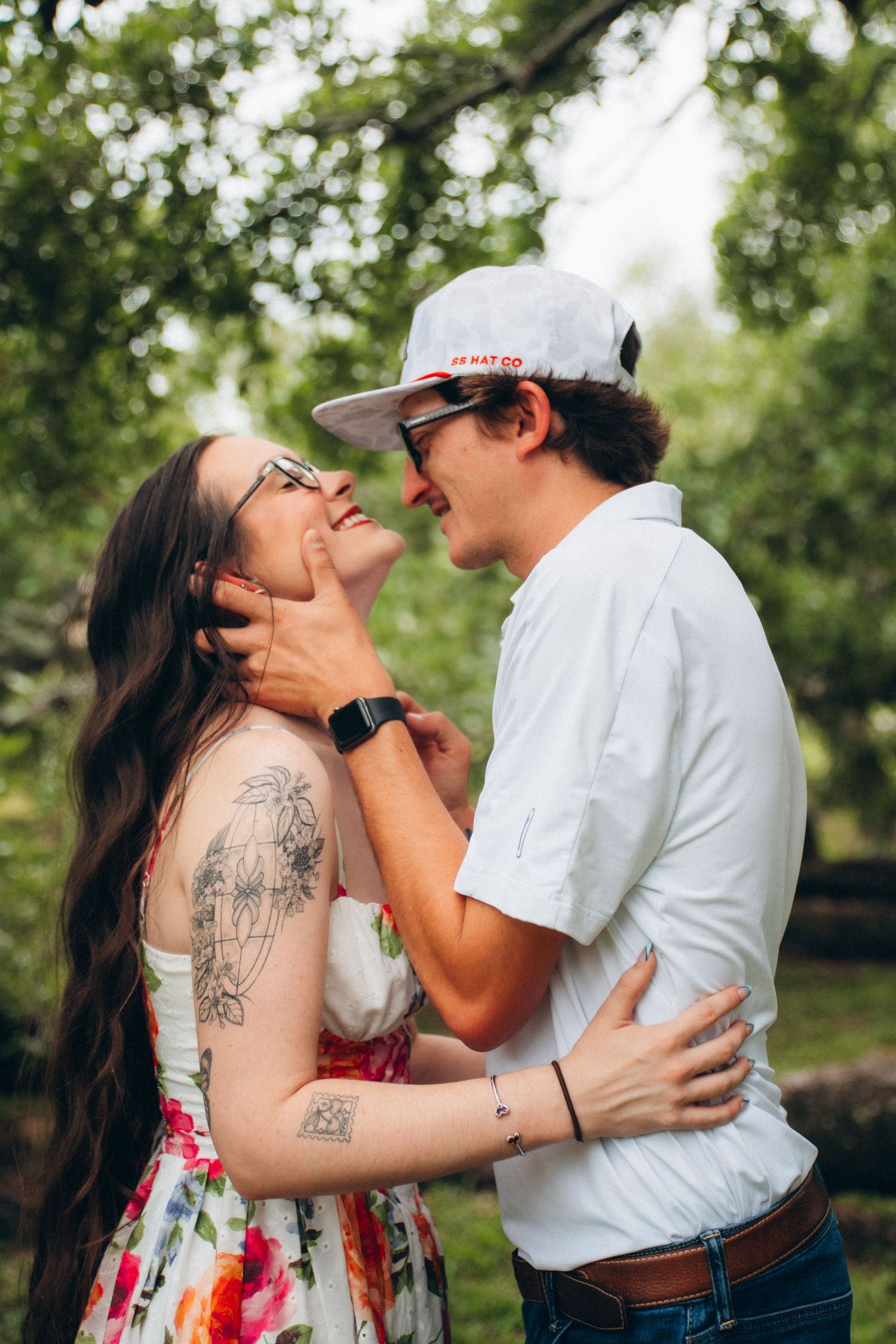 Couple sharing a tender moment in a lush green park, woman in floral dress with tattoos, man in white cap.