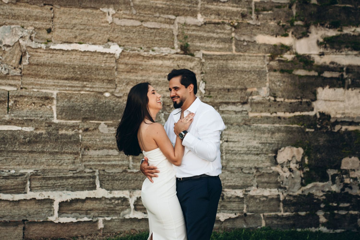 A bride and groom share a romantic moment laughing together against a rustic stone wall outdoors.