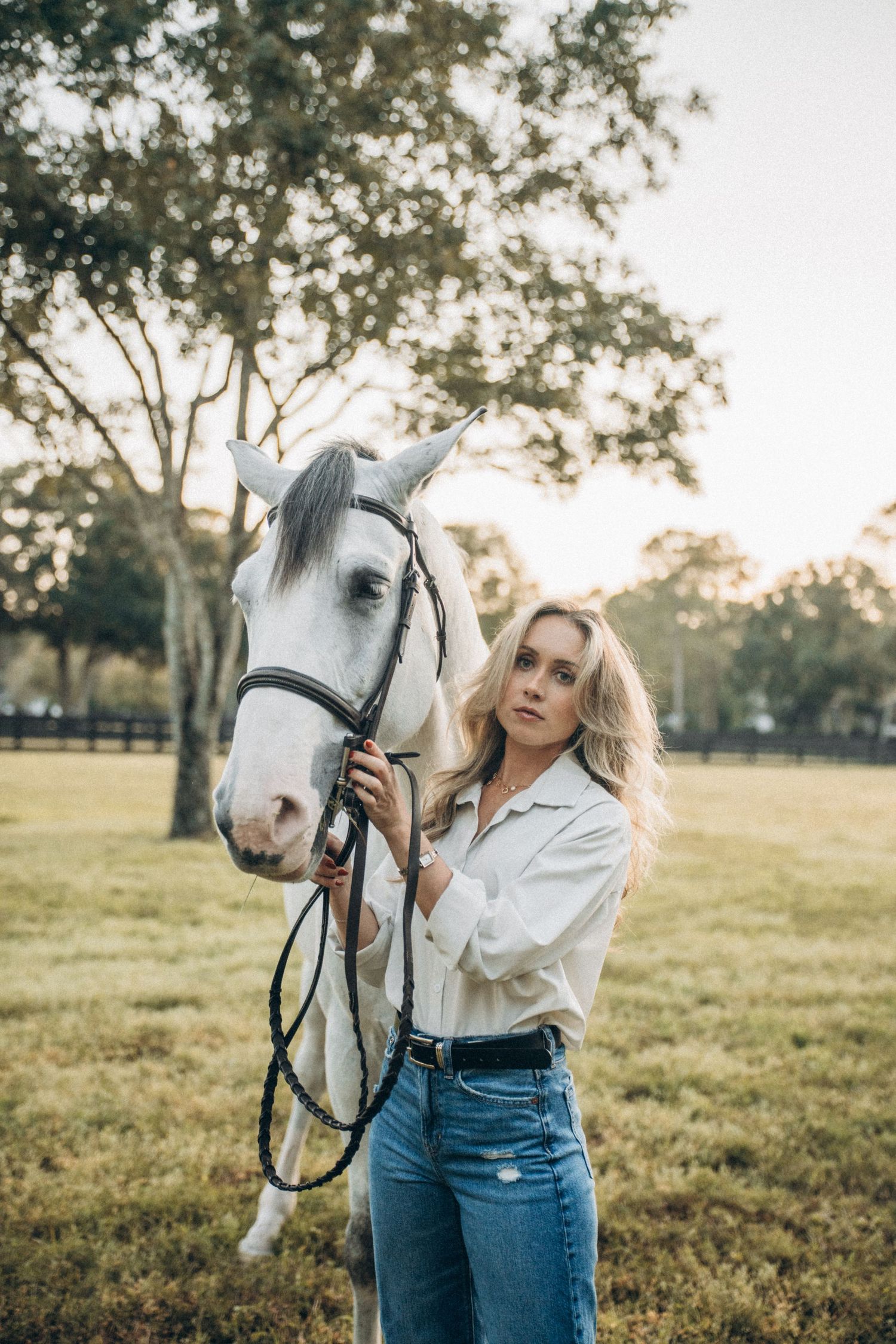 A white horse and person pose together in a pastoral field setting during golden hour.