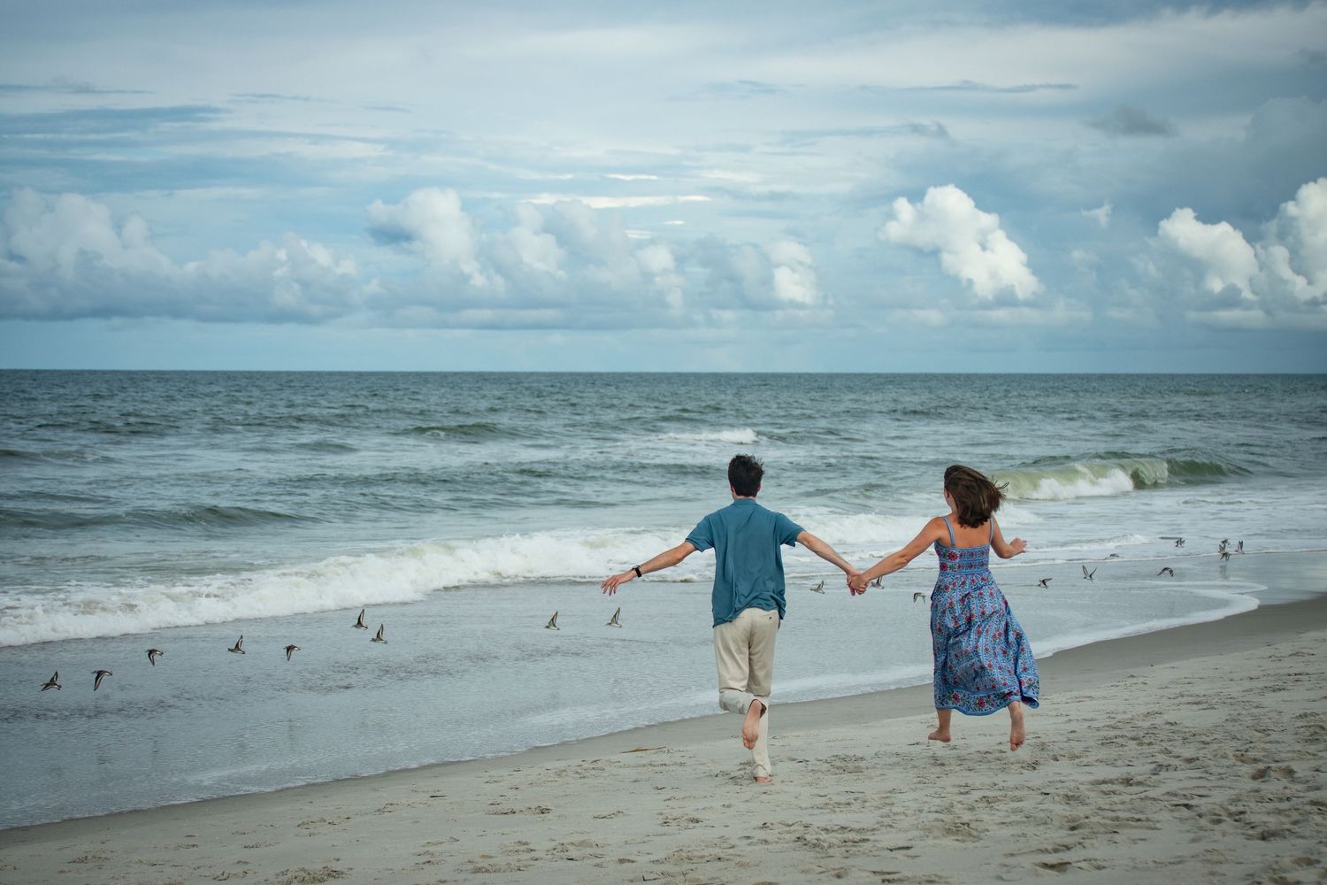 People walking along a serene beach shoreline with waves and cloudy skies in the background.