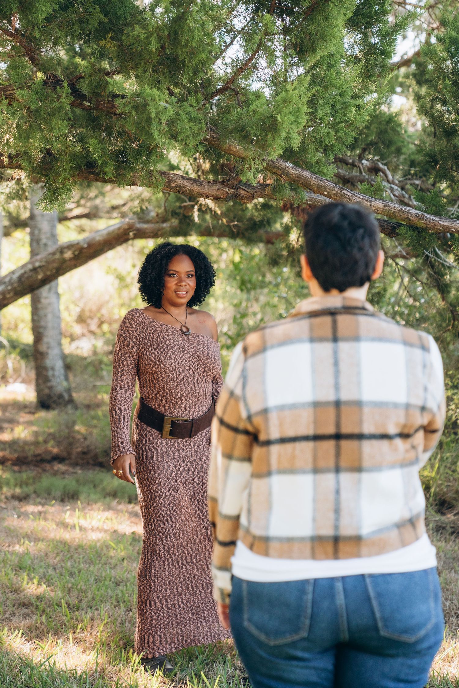 A couple stands together outdoors in a natural setting wearing coordinated brown and plaid outfits.