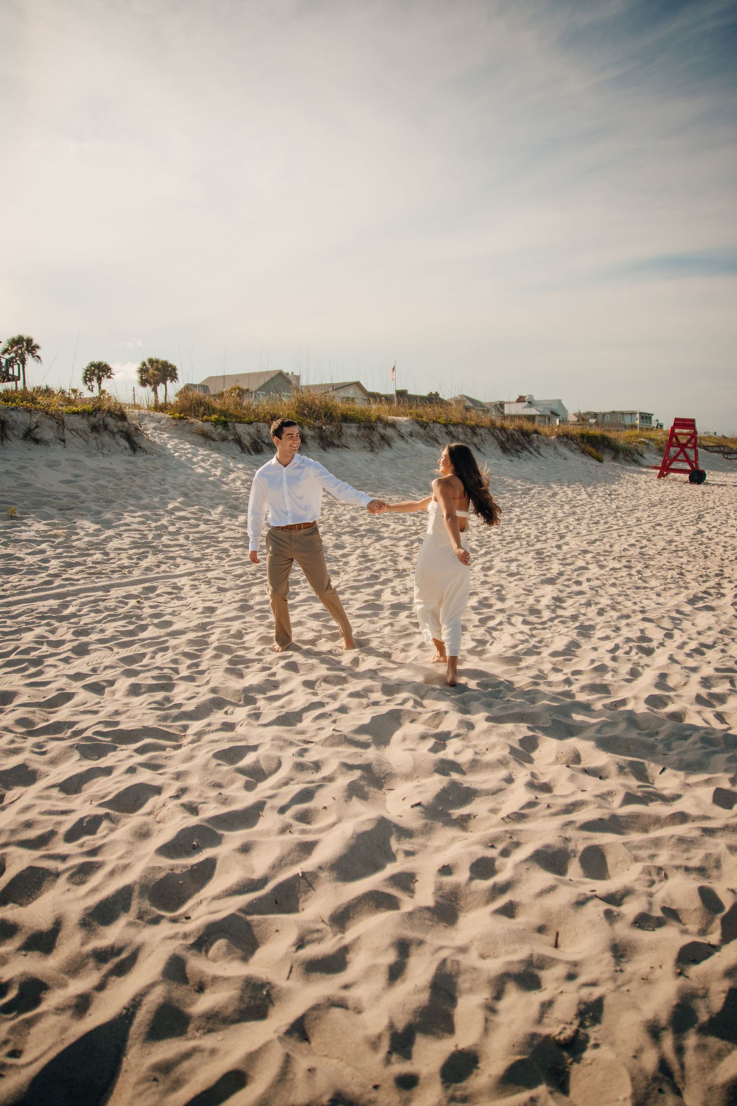 A couple walks hand in hand across a pristine white sand beach with coastal homes in the background.