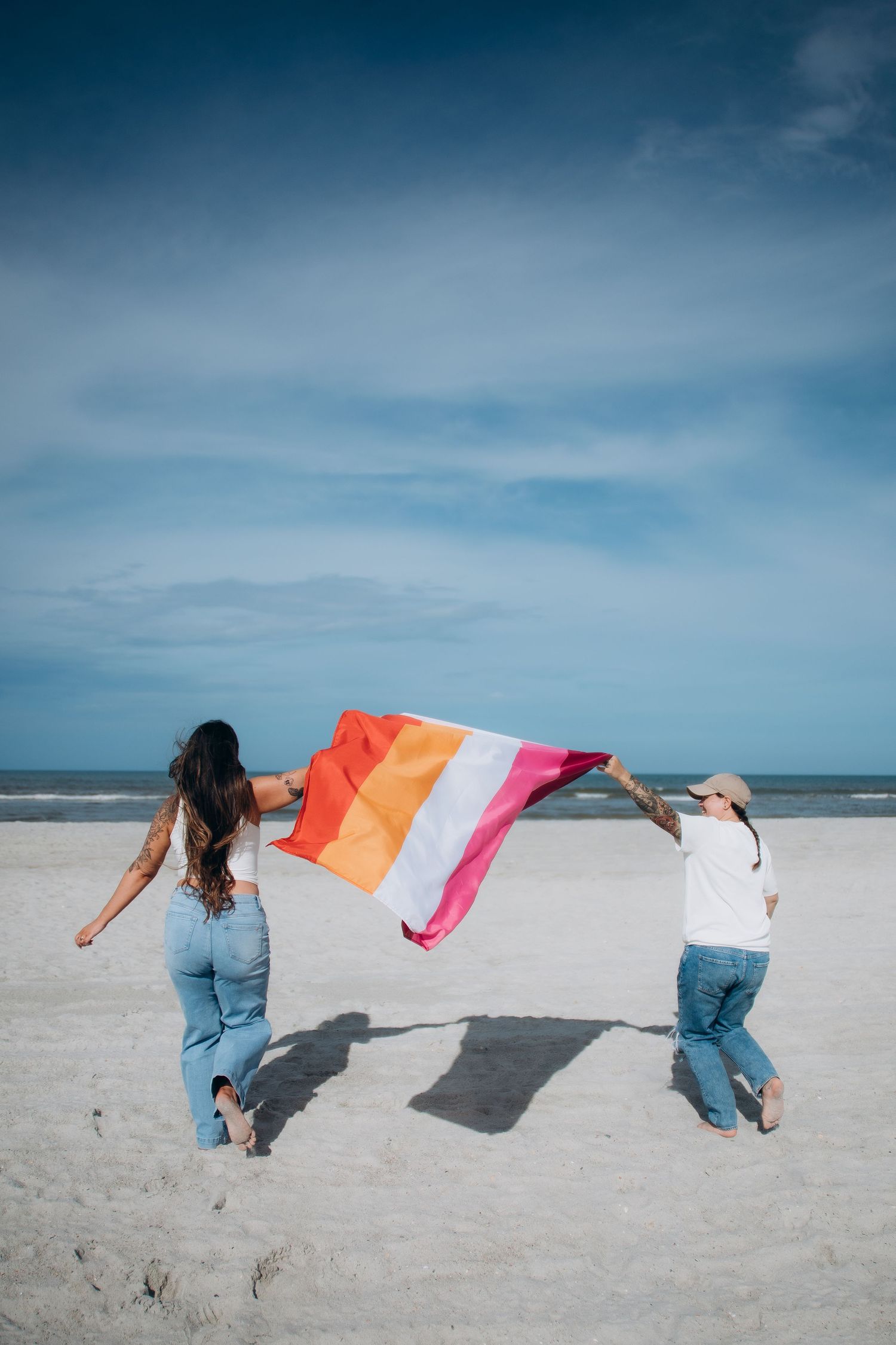 Two people running playfully on the beach with a colorful flag flowing behind them.
