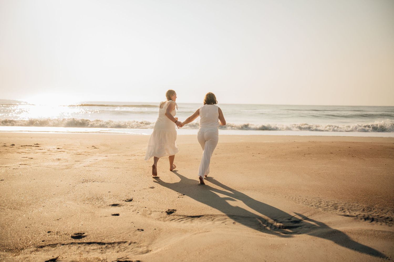 Two women in white dresses hold hands walking on a sunny beach toward ocean waves.