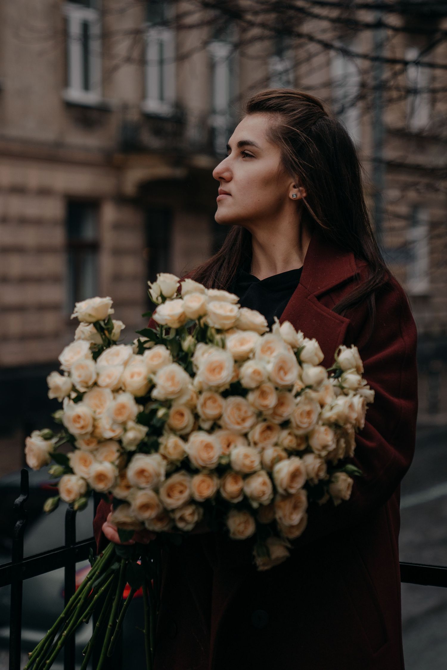 Young woman in burgundy coat holding bouquet of cream roses on a city street.