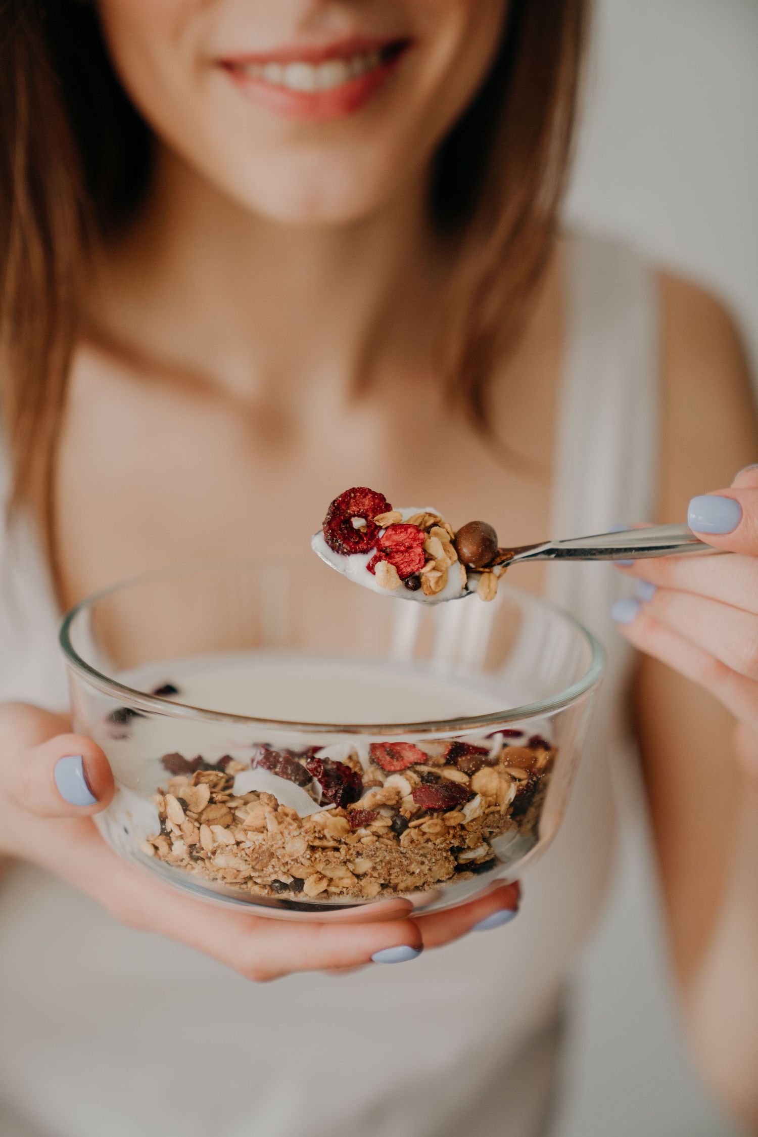 A smiling woman holding a glass bowl of granola with dried fruits and a spoonful close to the camera.