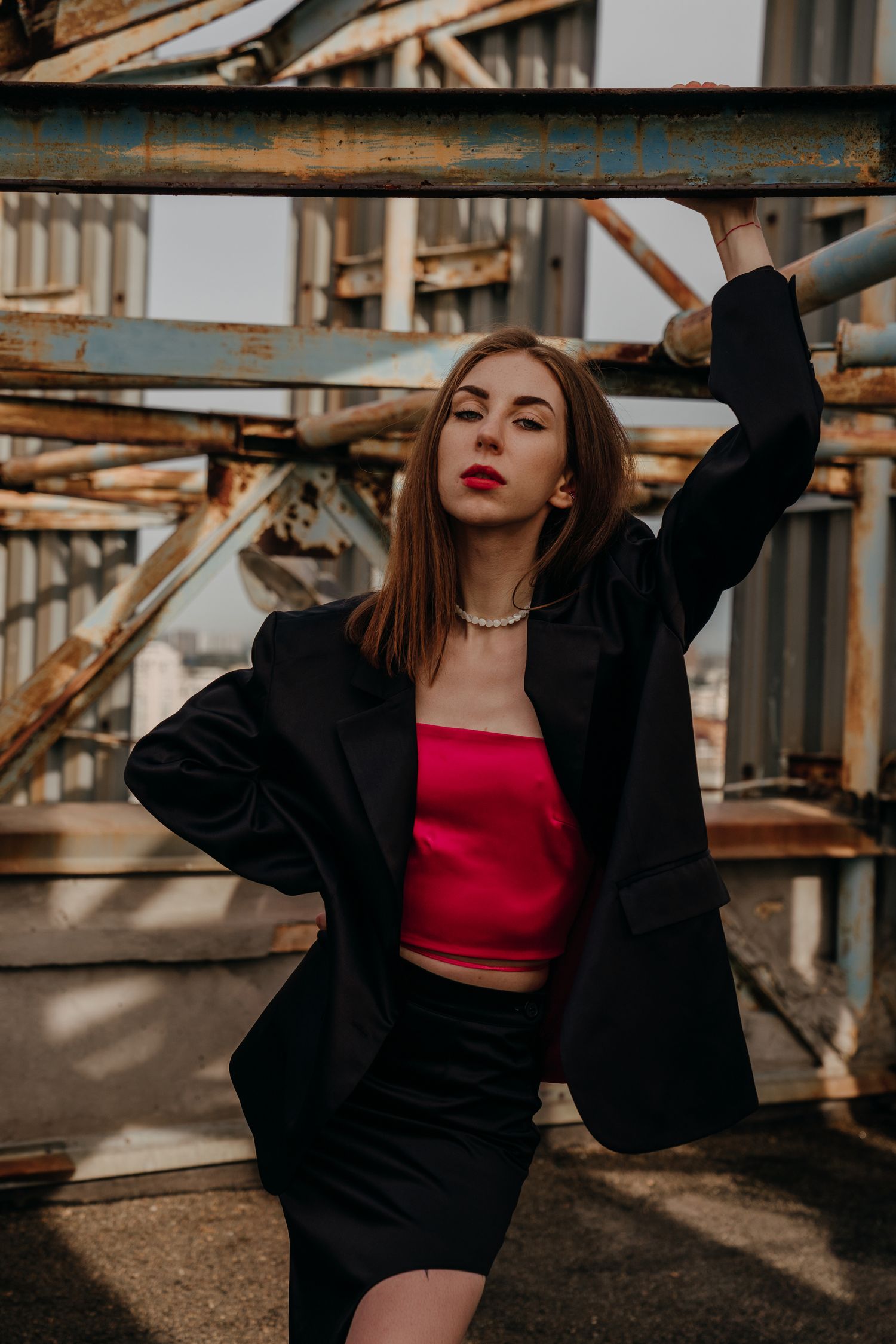 A young woman in a red crop top, black blazer and skirt poses confidently against rusty metal scaffolding outdoors.