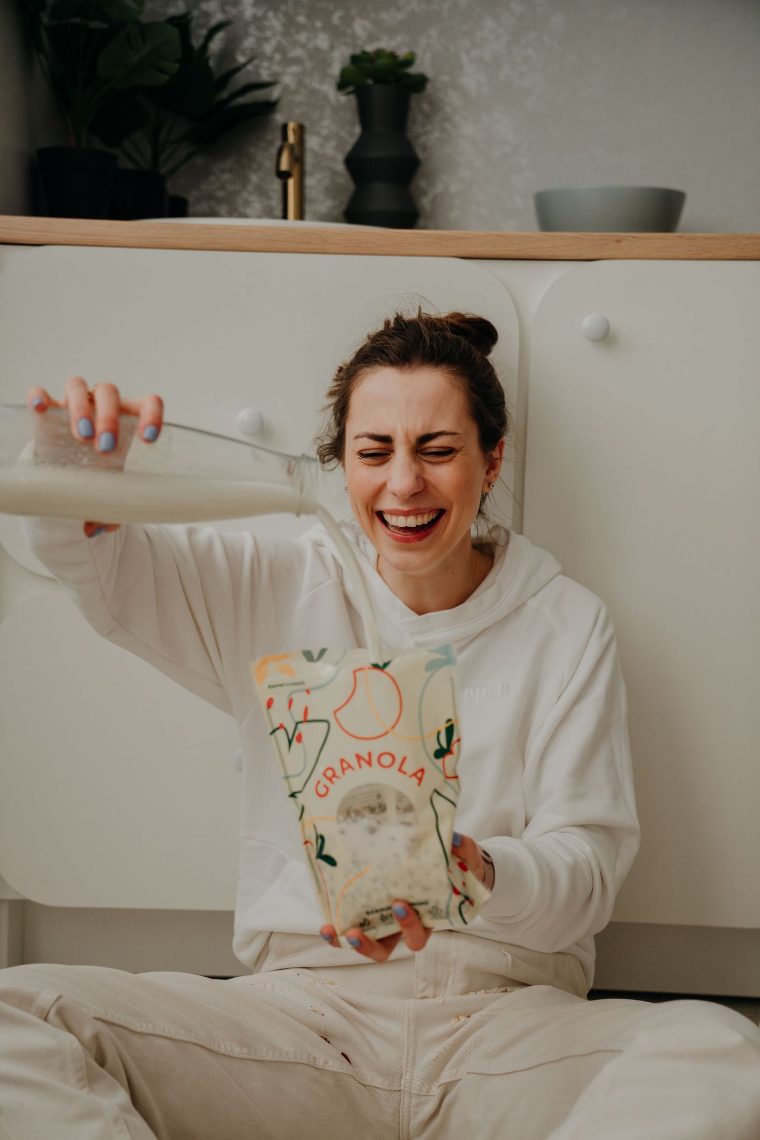 Woman in white hoodie laughing while pouring milk into a granola bag while sitting on a bed.
