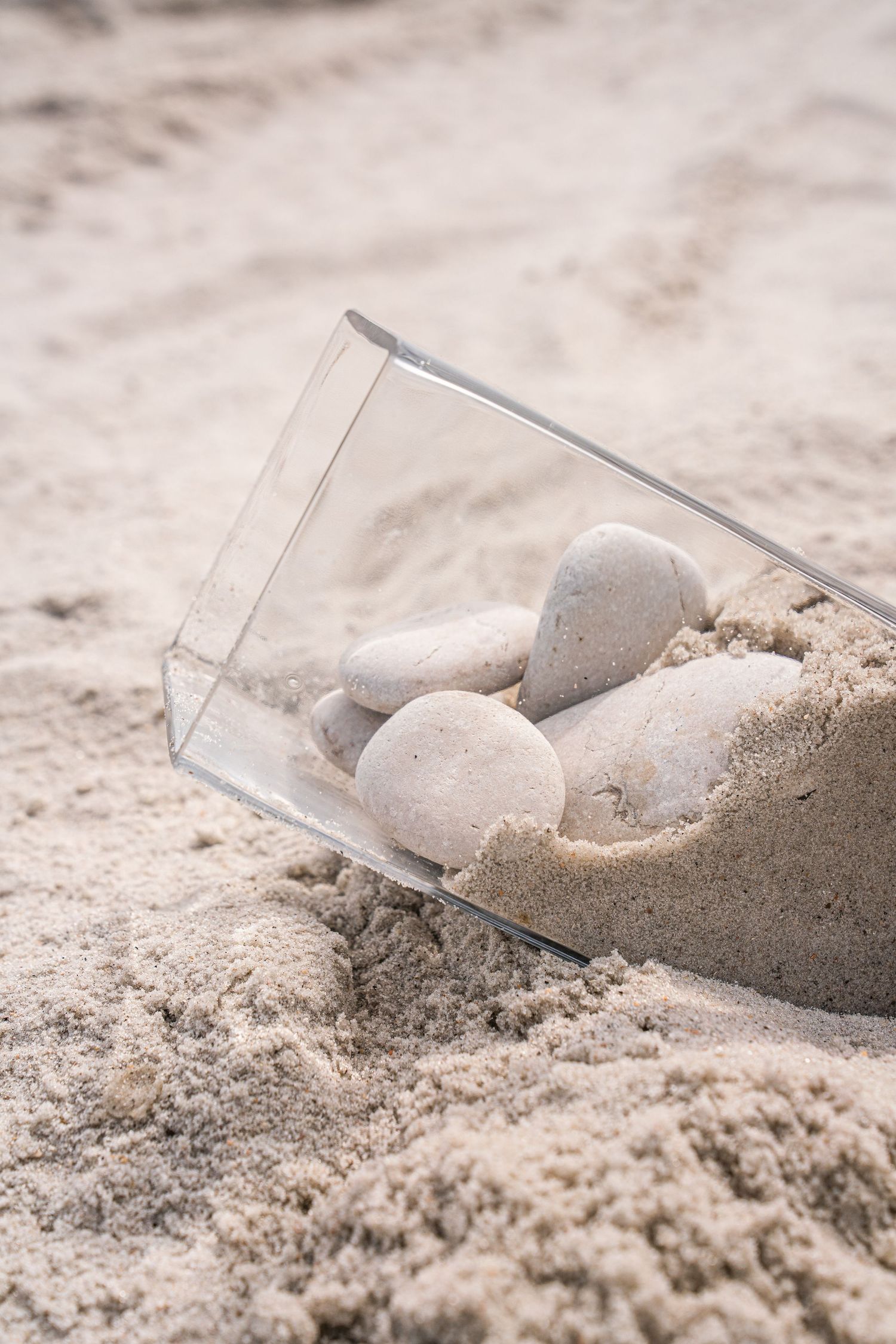Small smooth white stones inside a clear acrylic box partially buried in fine beach sand.