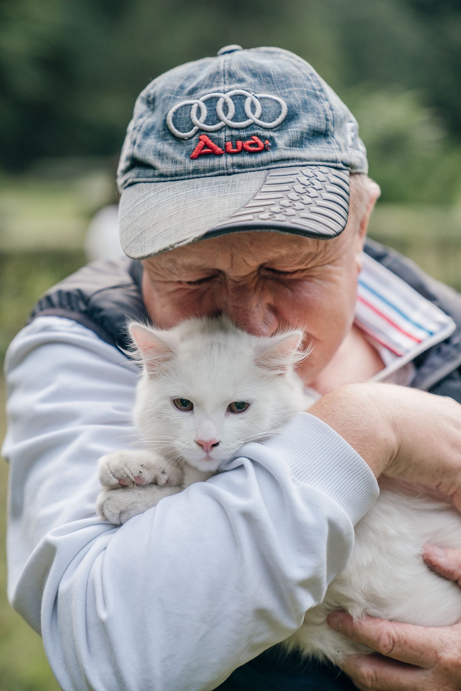 Elderly man wearing an Audi cap tenderly holding a white cat outdoors in a green natural setting.