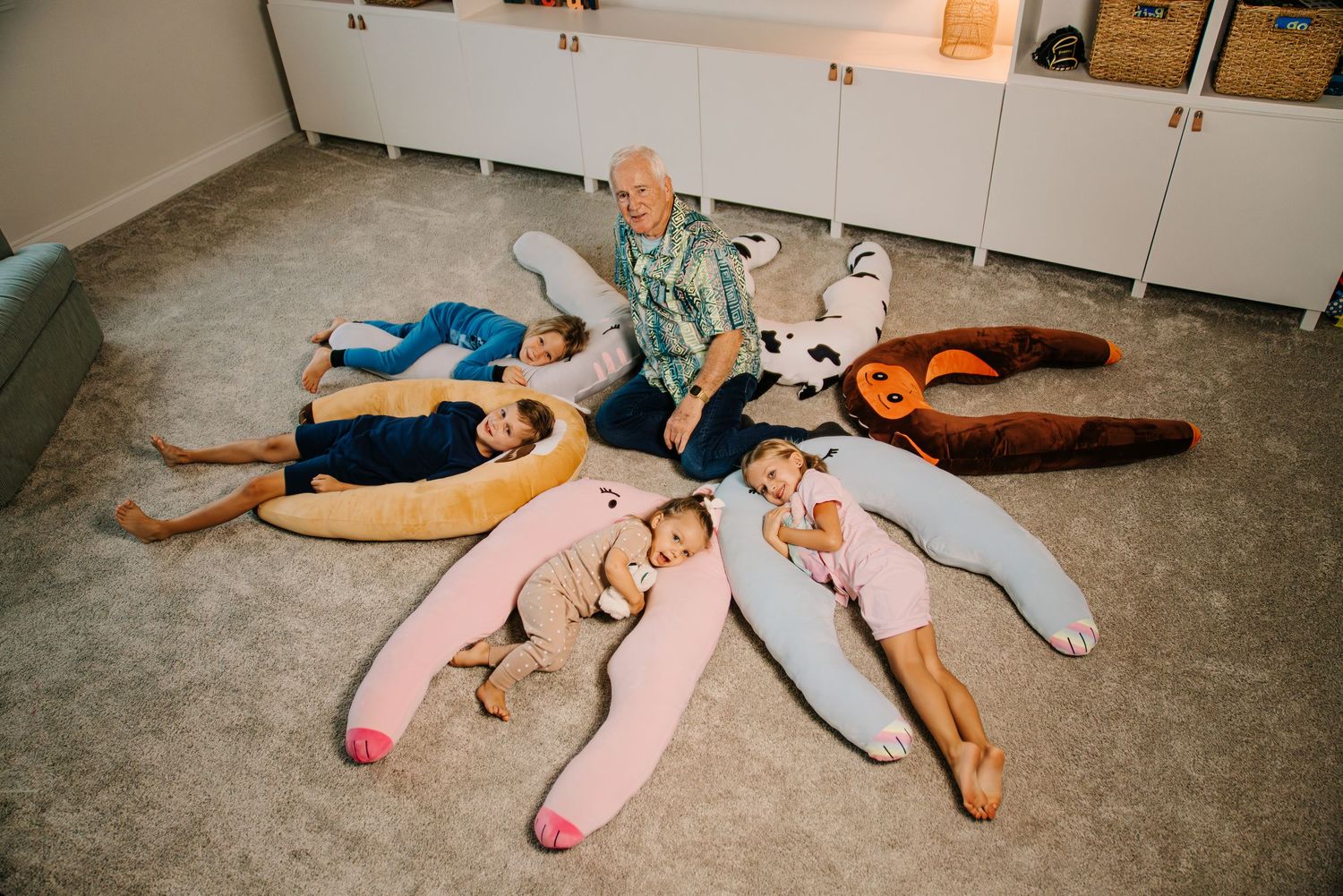 Children lying in a circle with giant stuffed animal toys while an adult sits in the center on a carpeted floor.