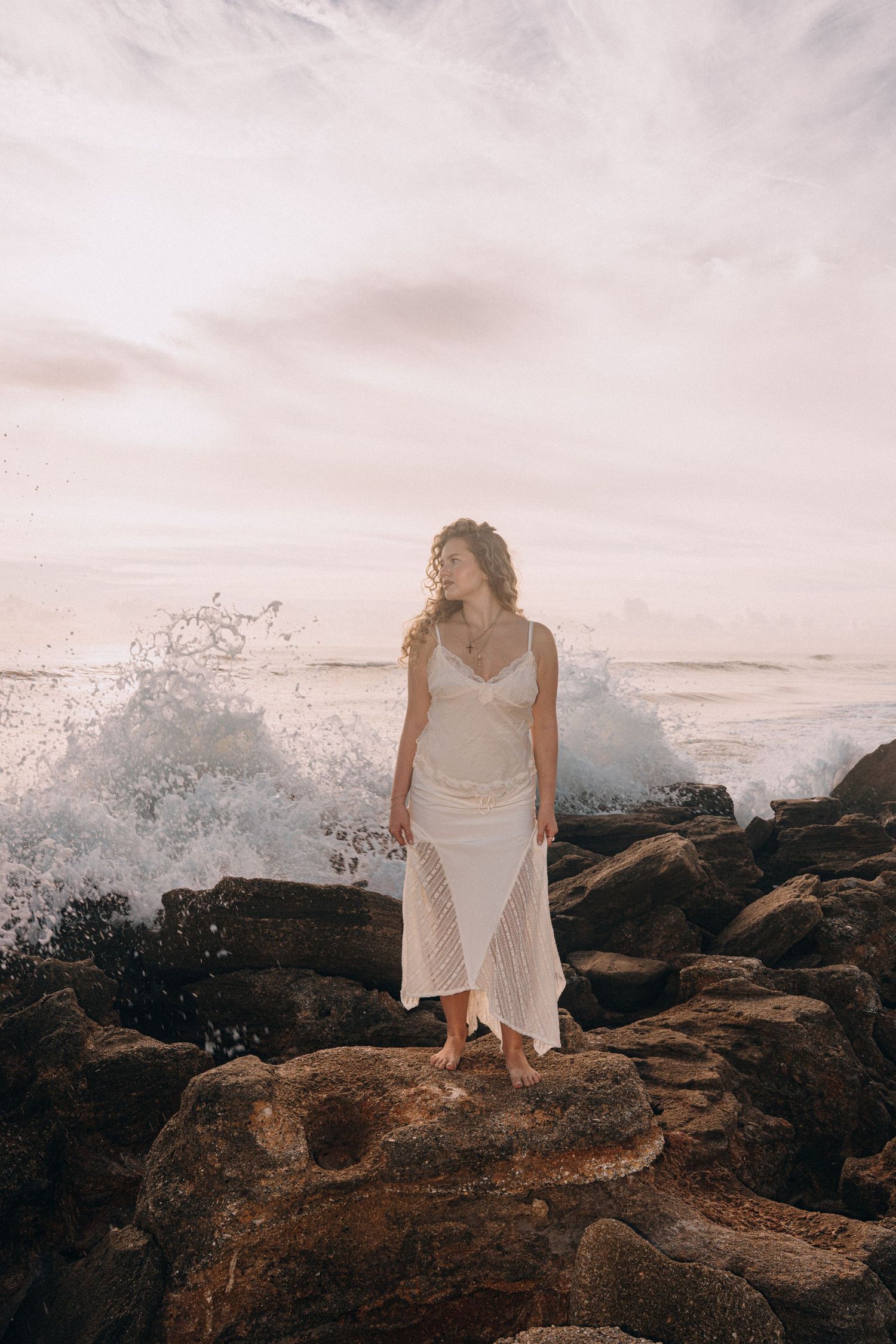 Woman in white dress standing on rocky shore with dramatic ocean waves crashing behind her at sunset.
