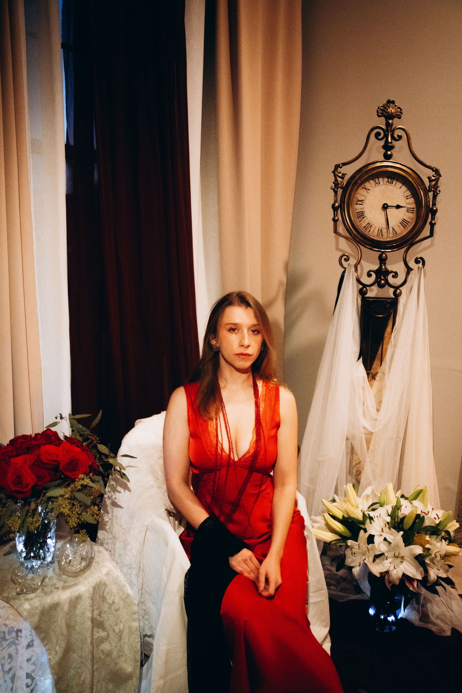 Young woman in red dress seated among flowers and vintage clock decor.