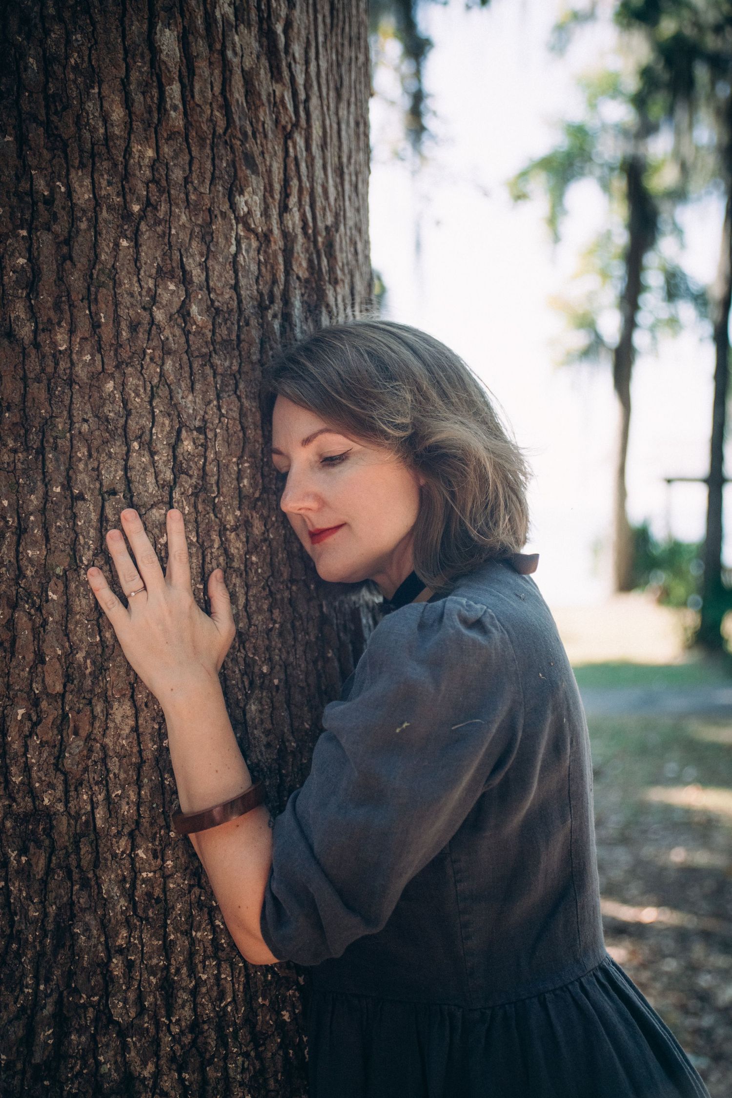 A person in dark clothing leans against a large tree trunk in a moody forest setting.