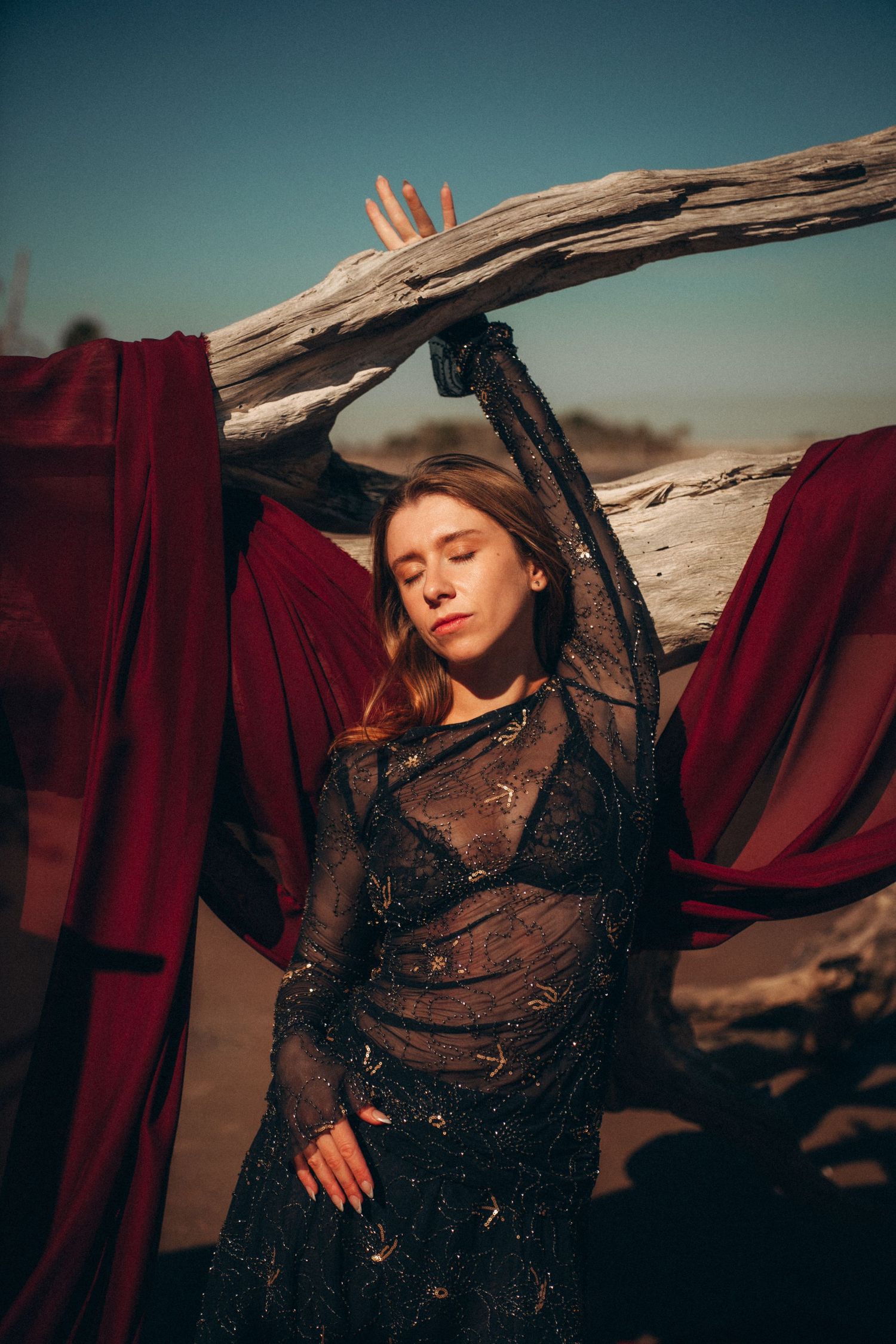 A dramatic fashion portrait in a desert setting with flowing red fabric and dark lace against weathered driftwood.
