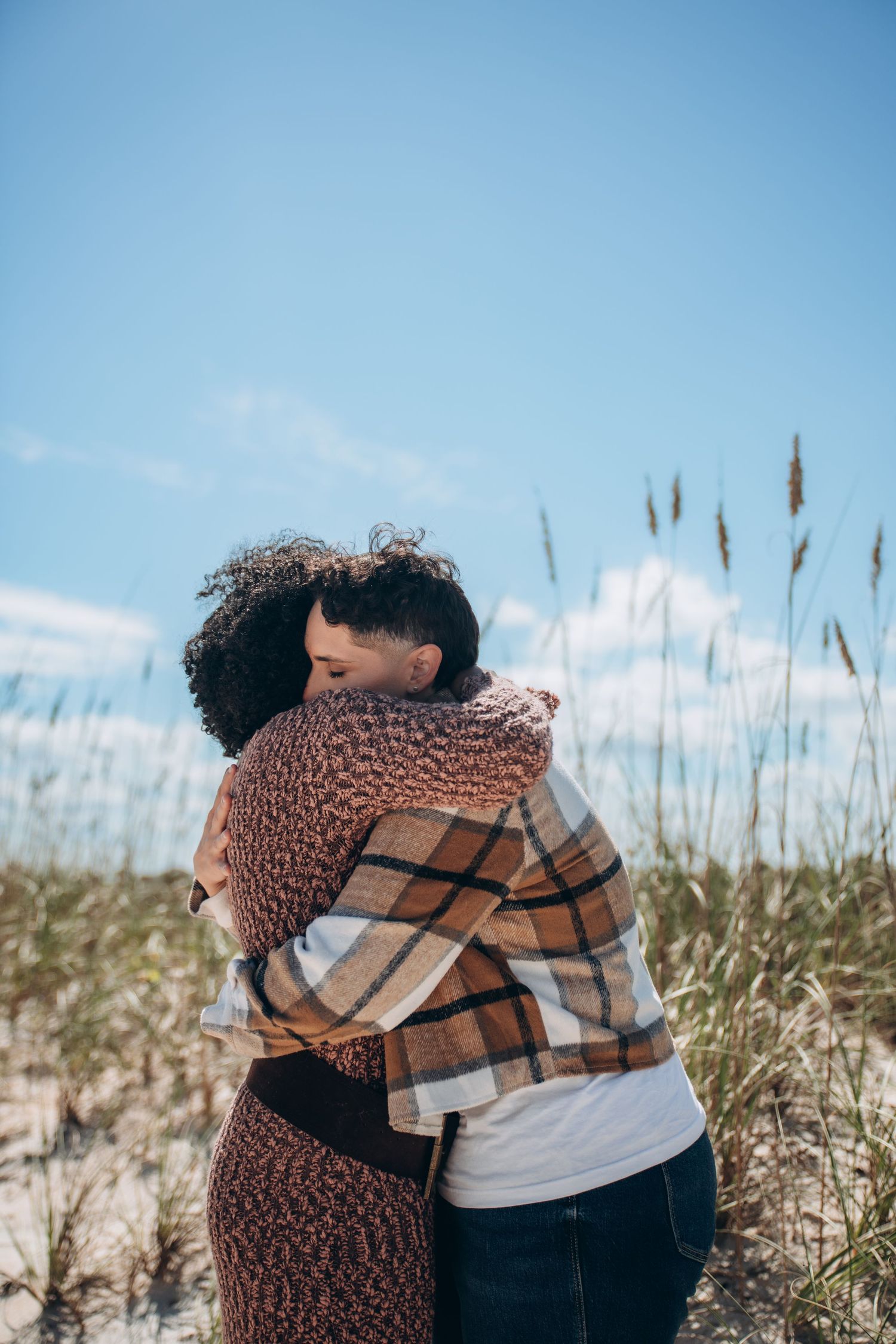 Two people share a warm embrace among tall grass and sand dunes against a bright blue sky.