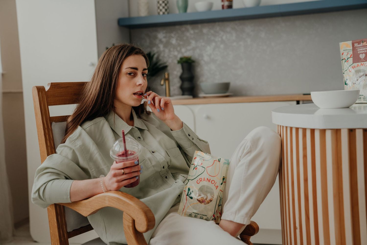 Young woman sitting in chair, sipping a drink through a straw while holding a snack bag in a cozy kitchen.