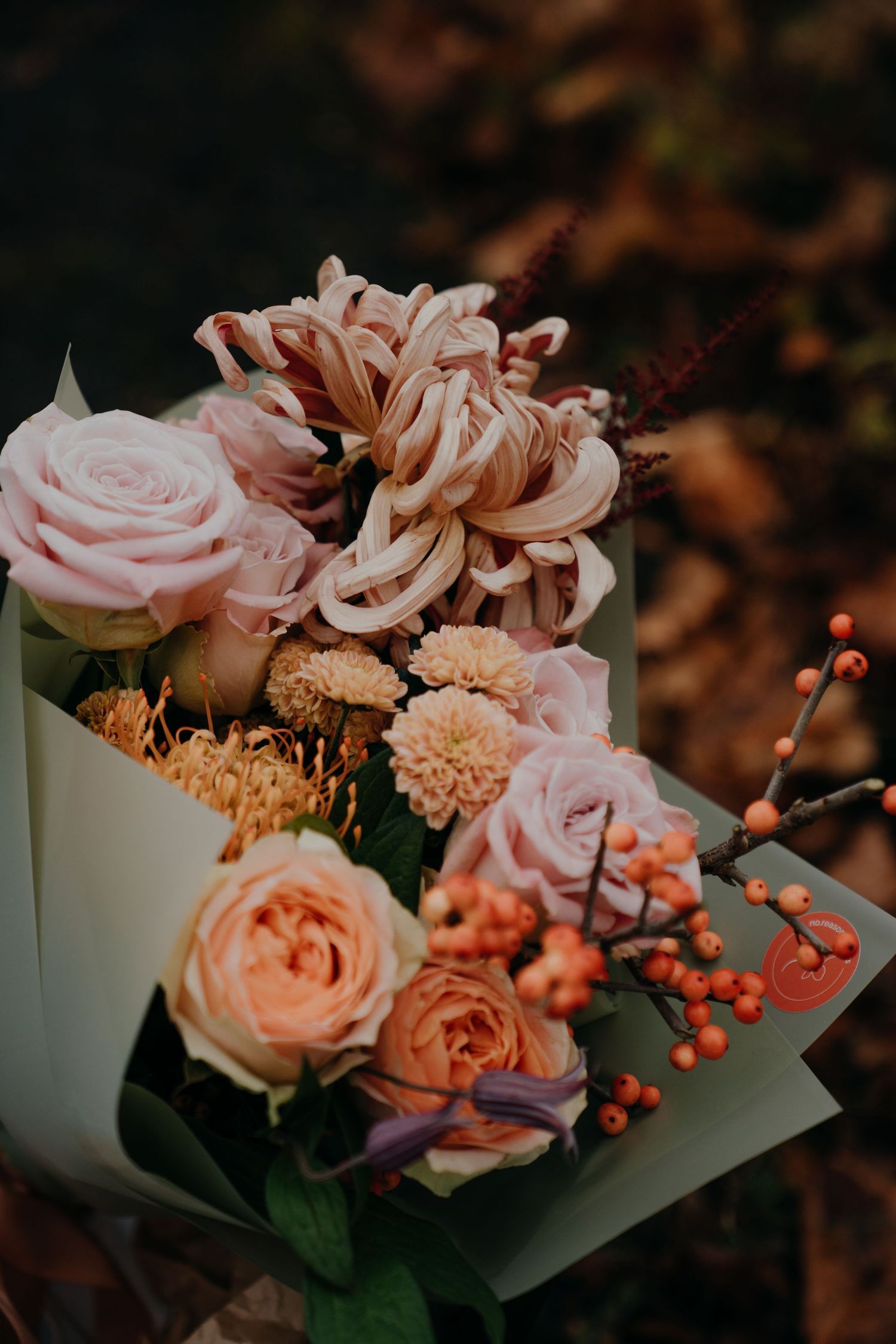 Autumn bouquet with pink roses, chrysanthemums, peach blooms, and orange berries wrapped in cream paper.