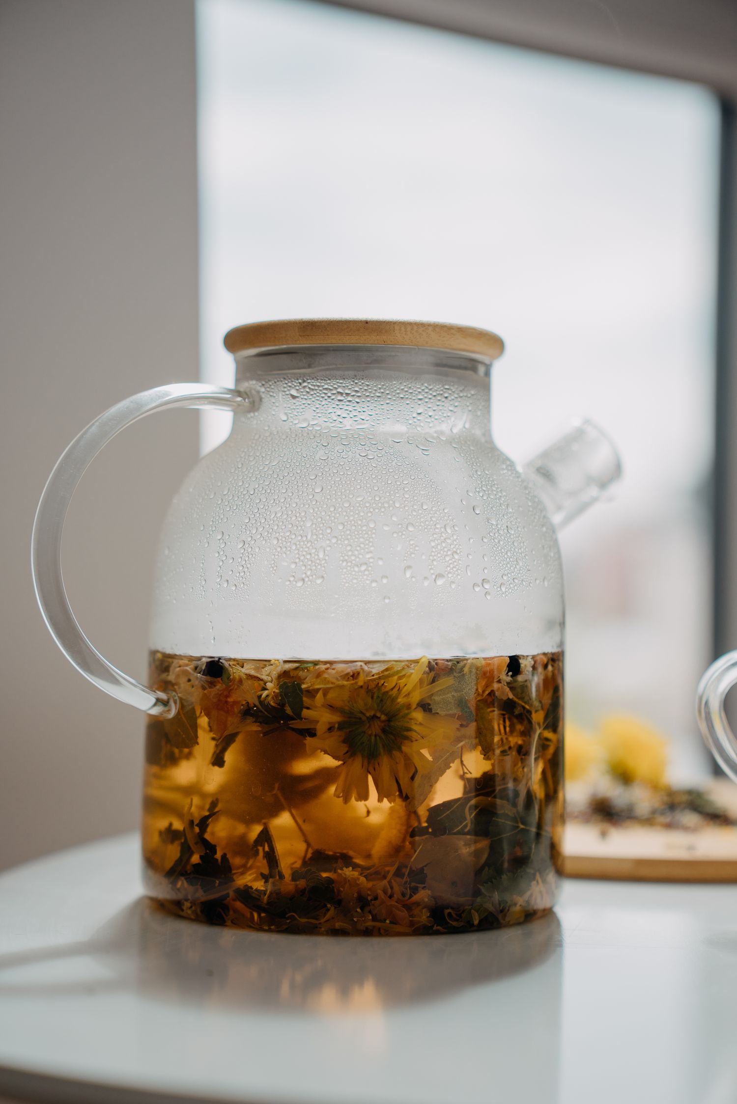 Glass teapot with bamboo lid filled with flowering herbal tea on a white surface.