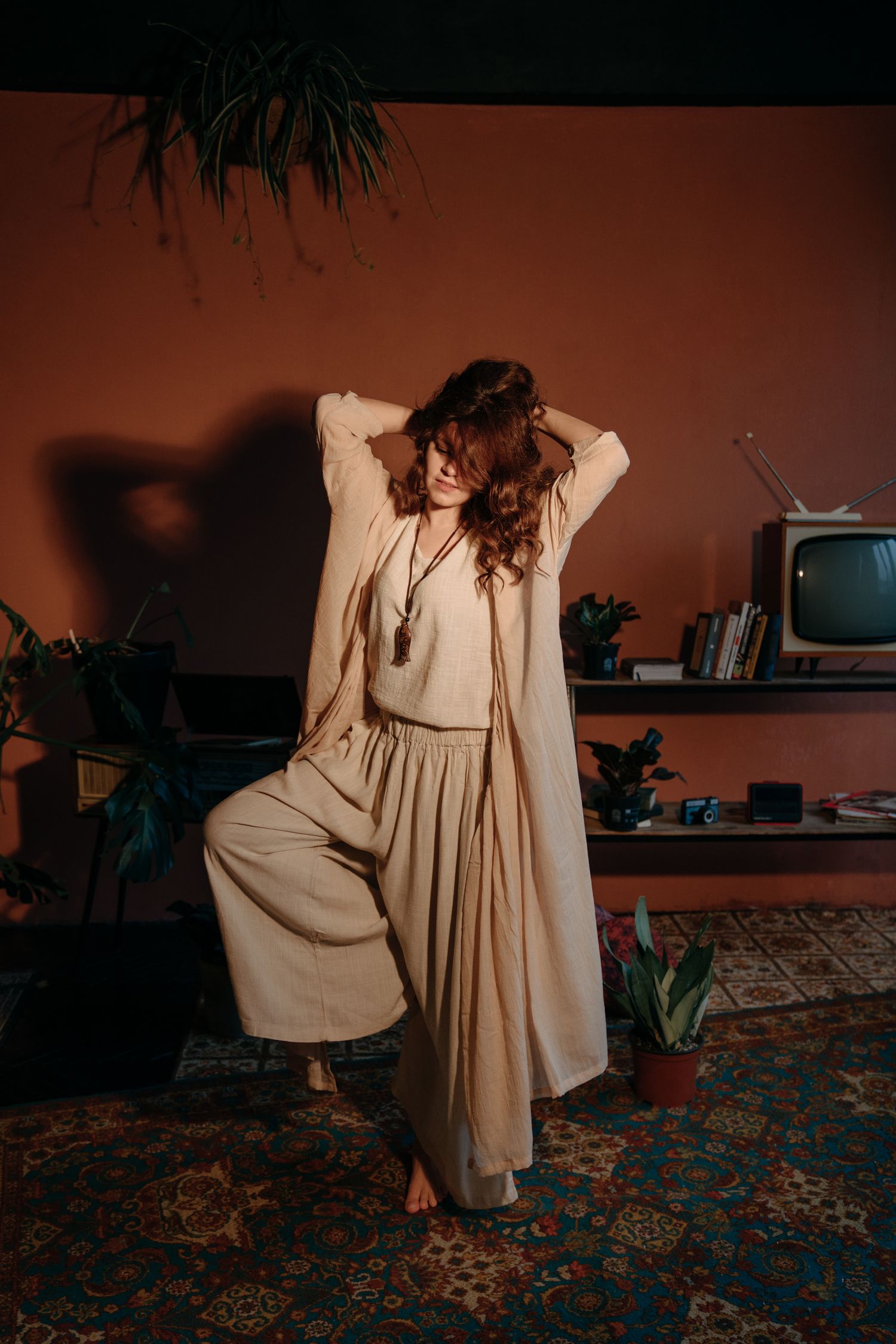 Woman in beige wide-leg pants and long cardigan poses barefoot in a retro-styled room with plants and vintage TV.