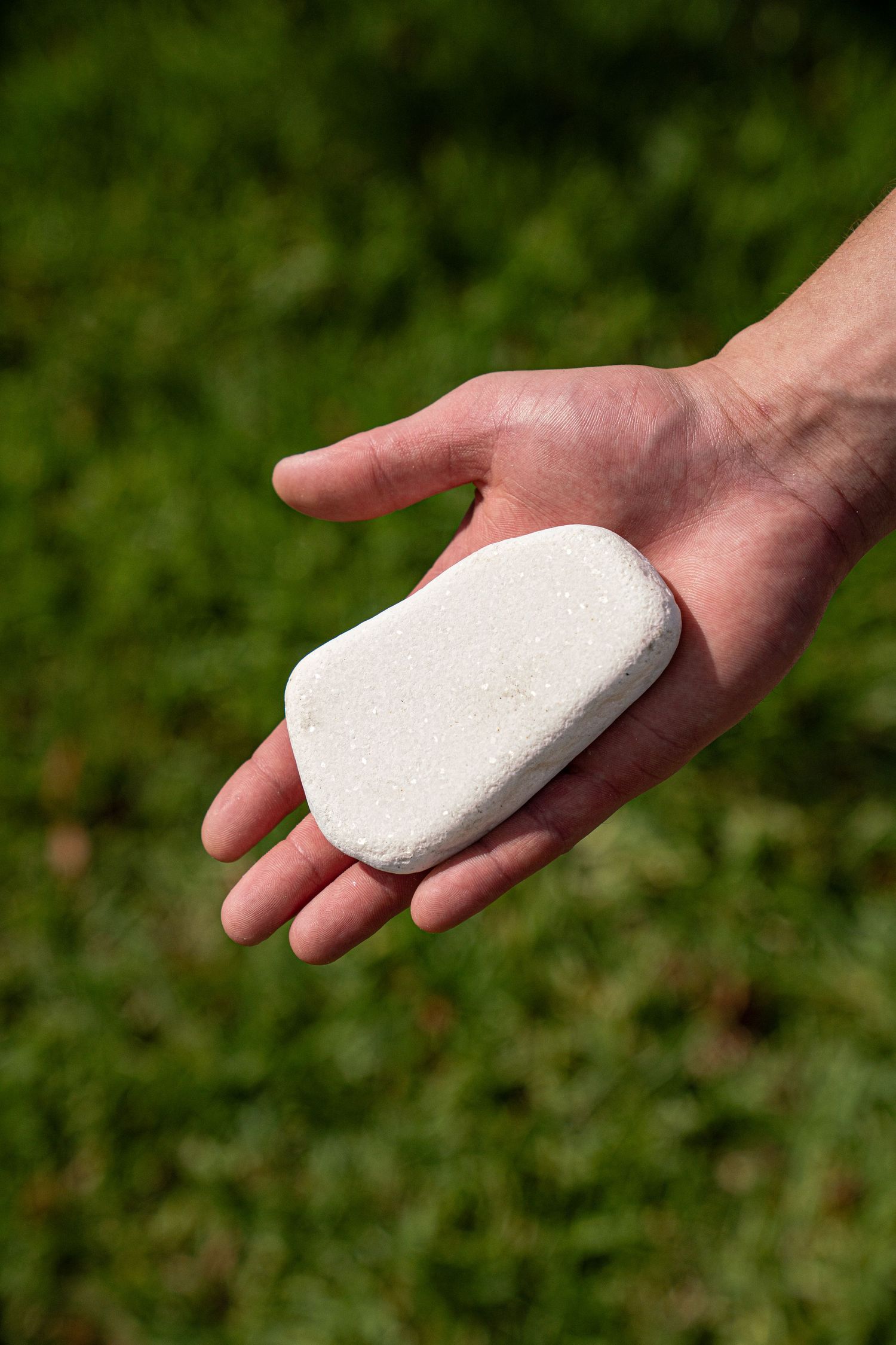 Hand holding a single smooth flat white stone against a blurred green grass background outdoors.