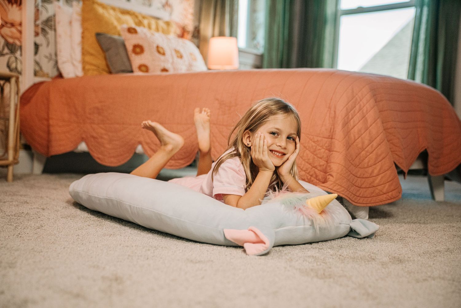 Young girl lying on carpet beside a large gray whale plush pillow in a cozy bedroom with orange bedding.