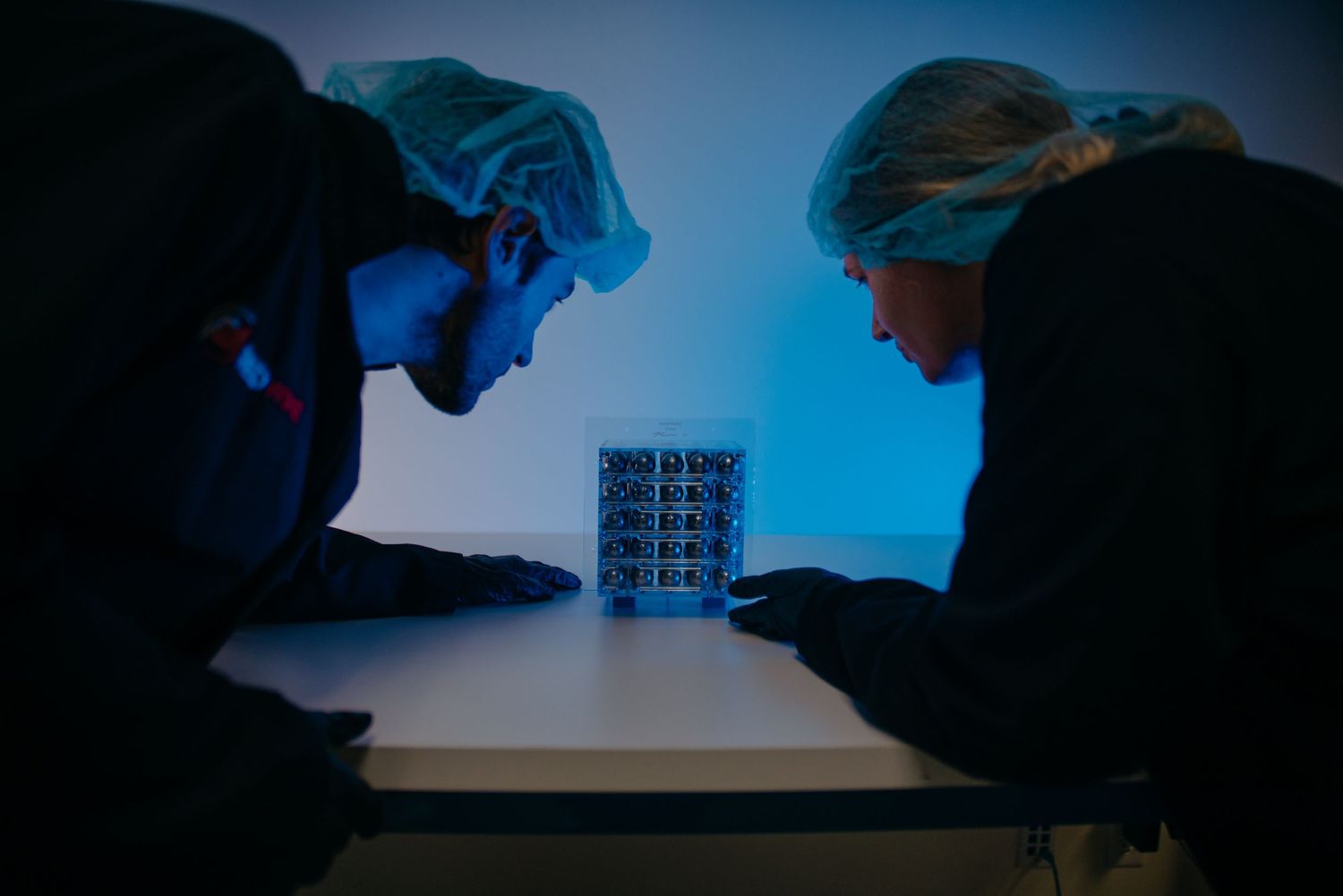 Two scientists in hairnets examine a microchip under blue laboratory lighting in a darkened cleanroom environment.