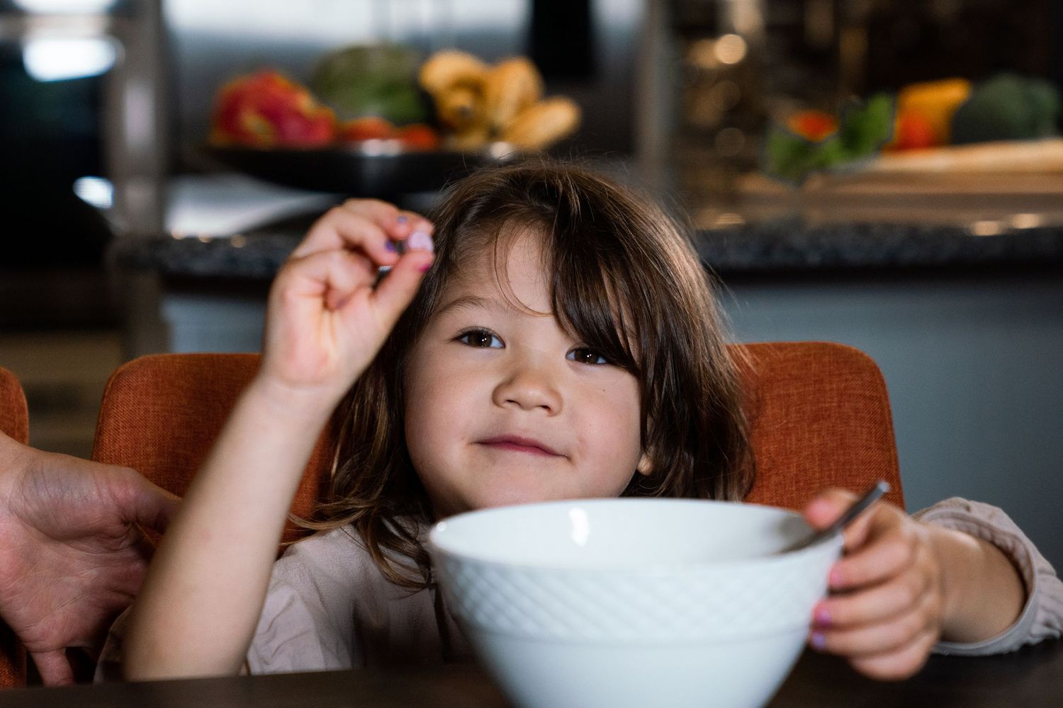 A smiling young girl sits at a dining table holding a spoon beside a white bowl, ready to eat a meal.
