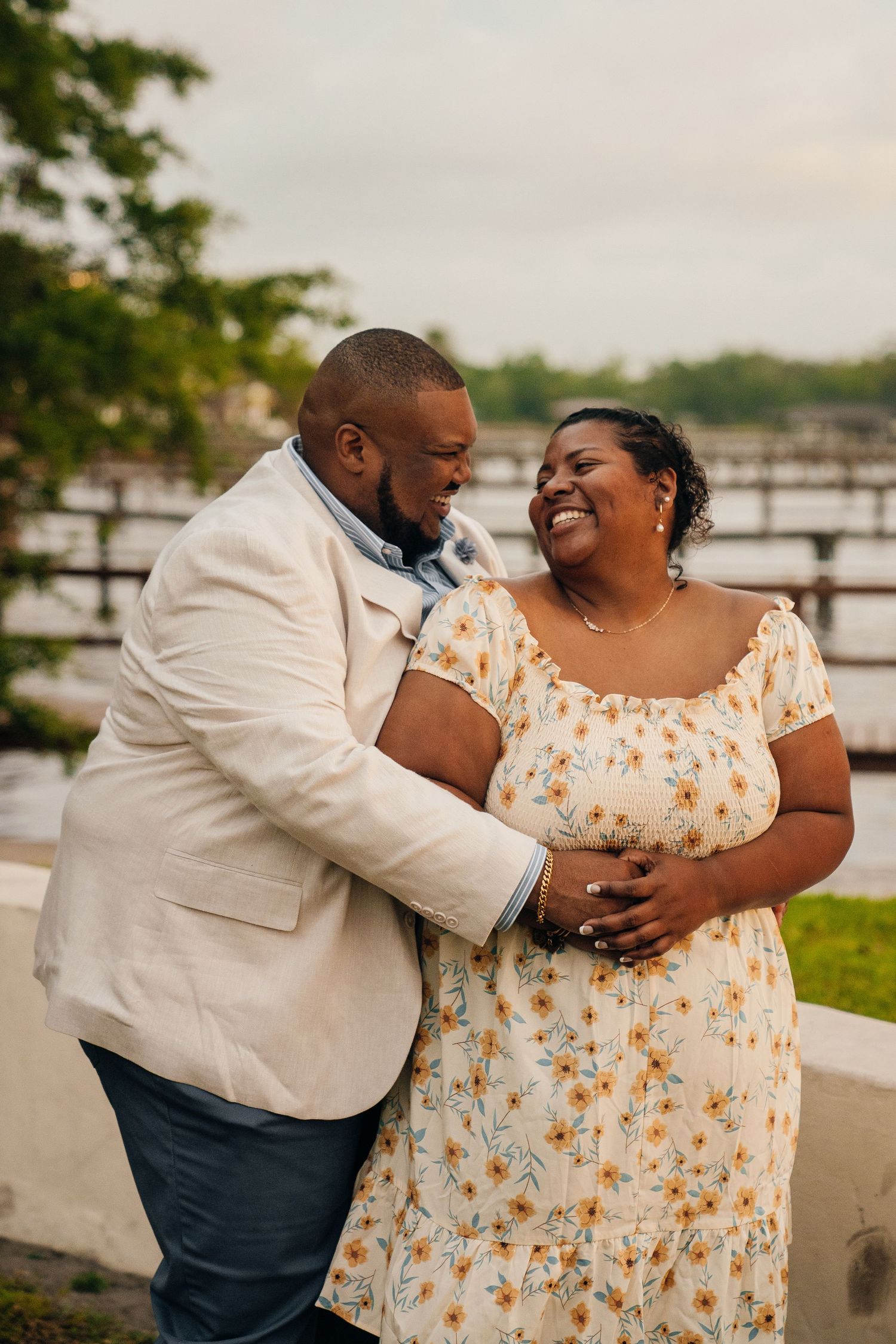 A joyful couple embraces outdoors, wearing a cream blazer and floral dress by the waterfront.
