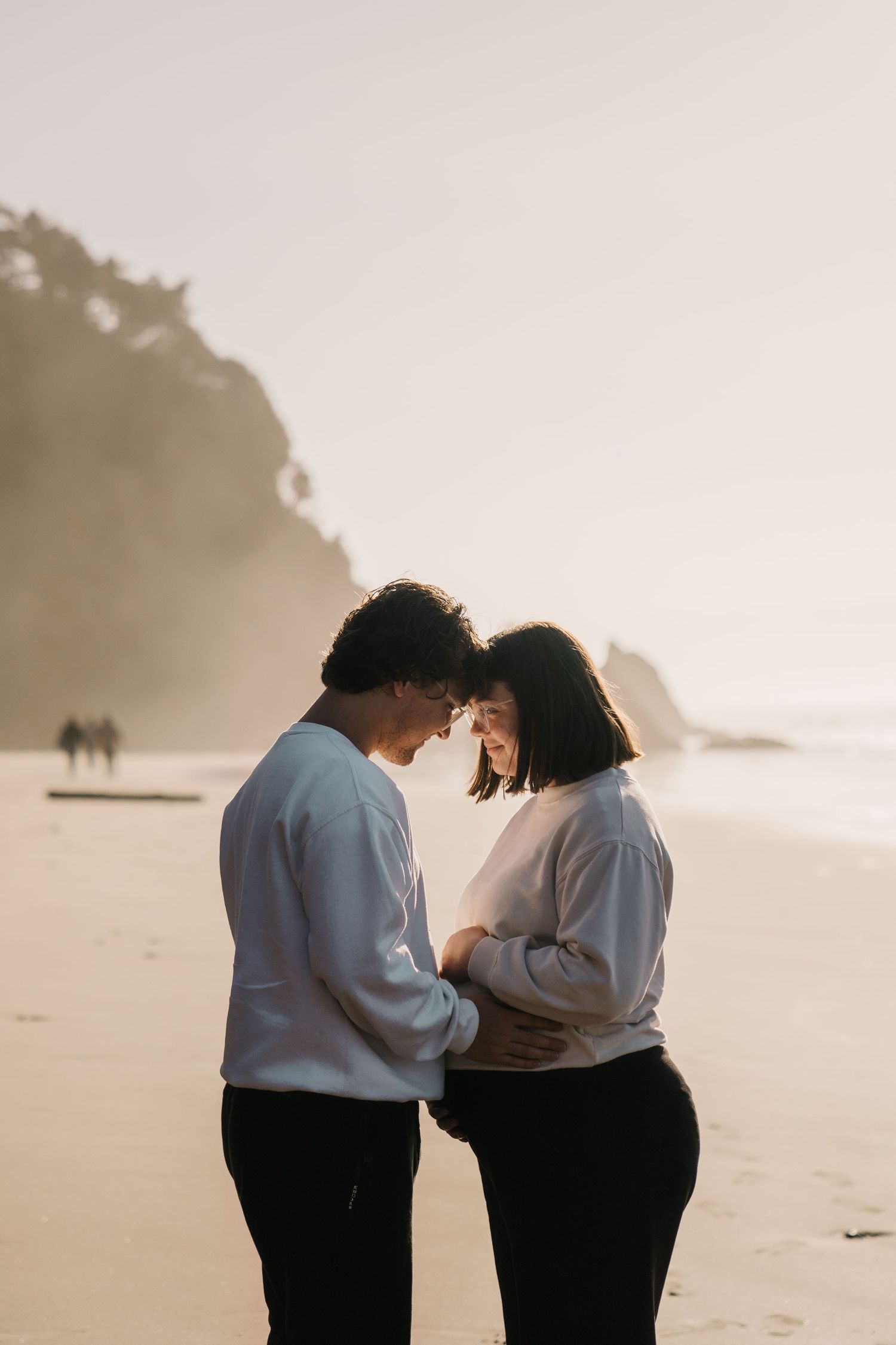 A couple embraces on a misty beach during sunset wearing white and black outfits.
