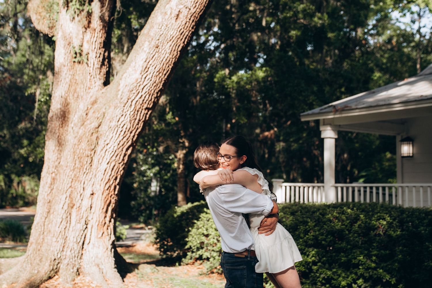 Couple shares playful moments under large oak trees near a white porch.