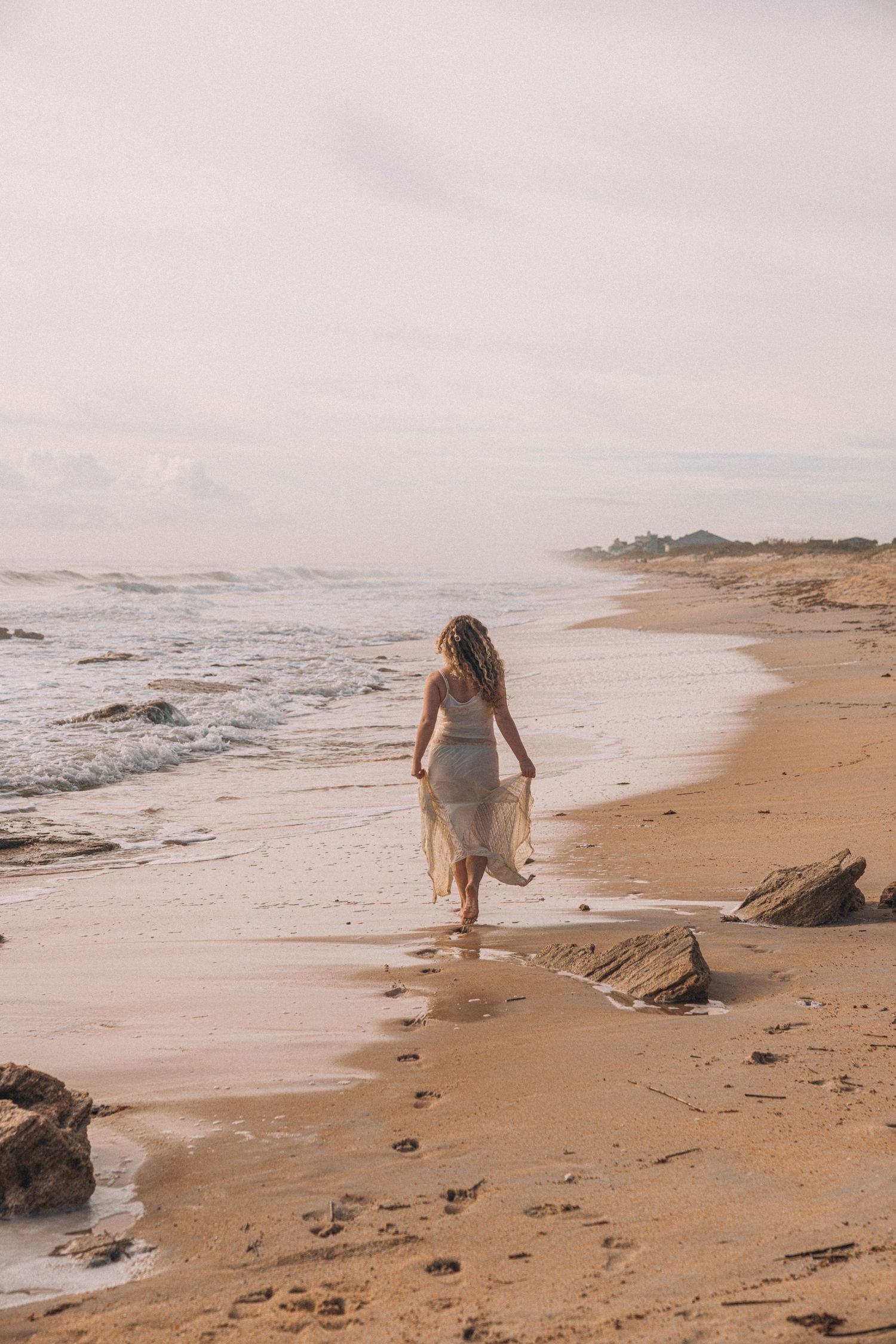 Woman in white dress walking along sandy beach shoreline on overcast day.