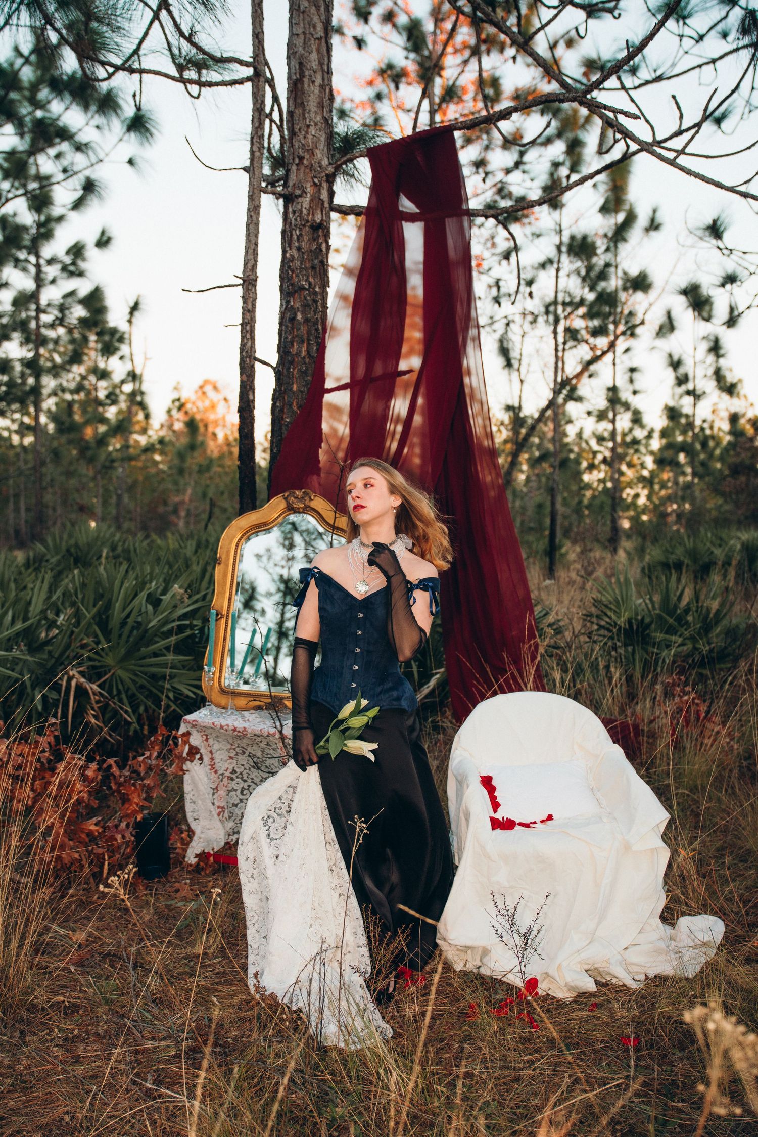 Elegant woman in black off-shoulder gown poses in forest setting with vintage mirror and red draping at dusk.